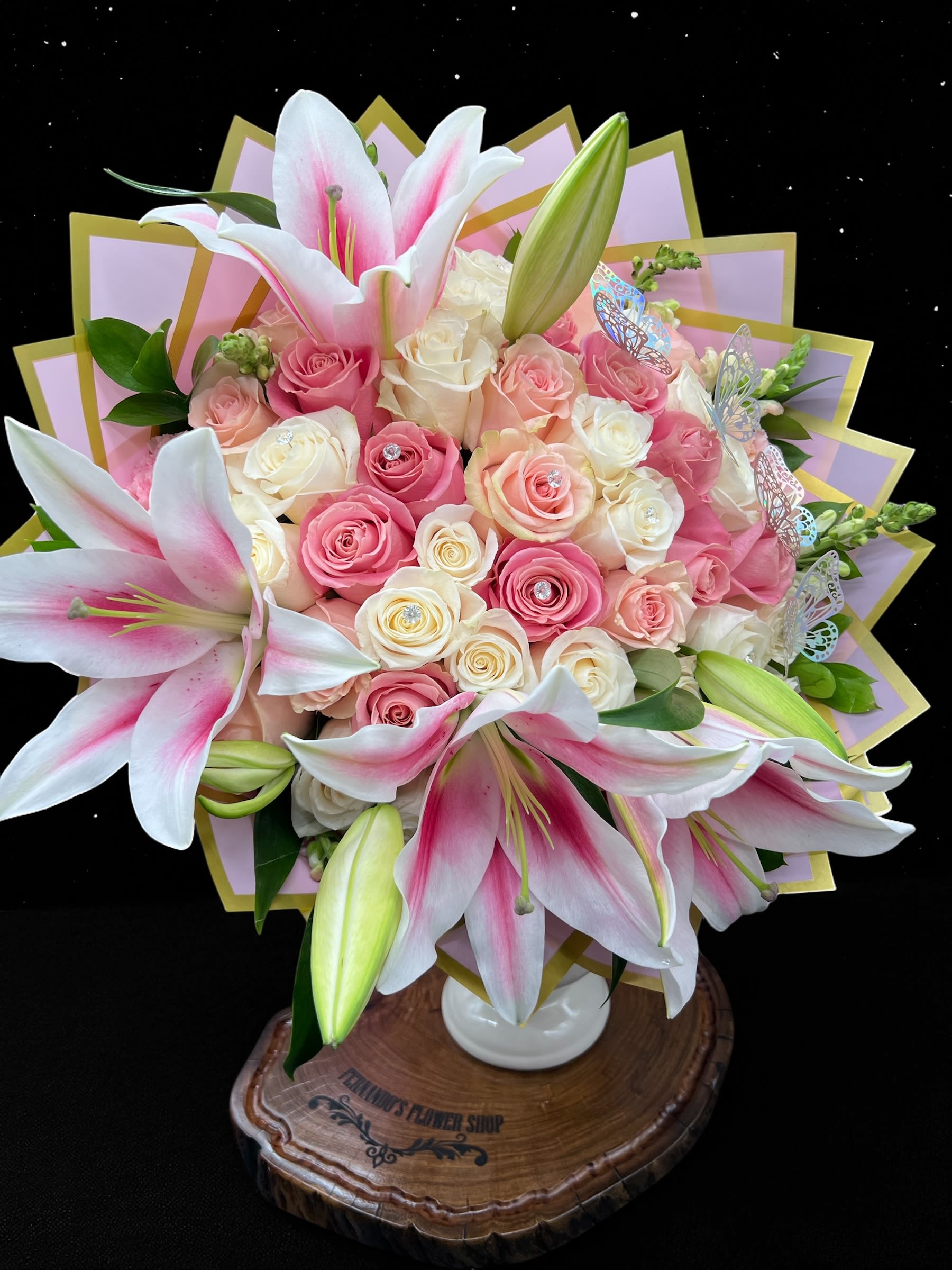 Pink lilies and cream and pink roses arranged in a white vase with decorative wrap and butterfly accents