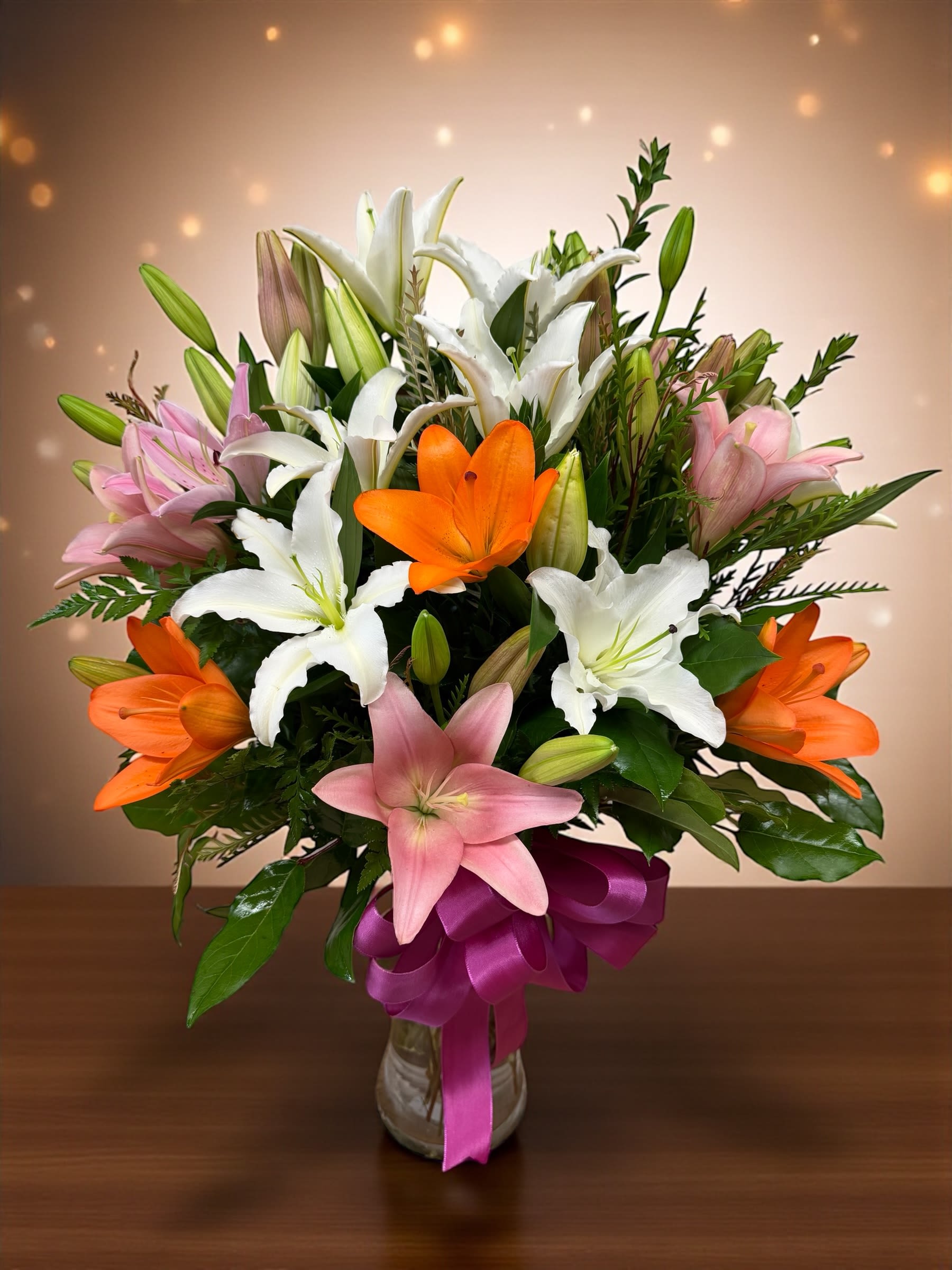 Bouquet of white, pink, and orange lilies in a glass vase with a purple ribbon