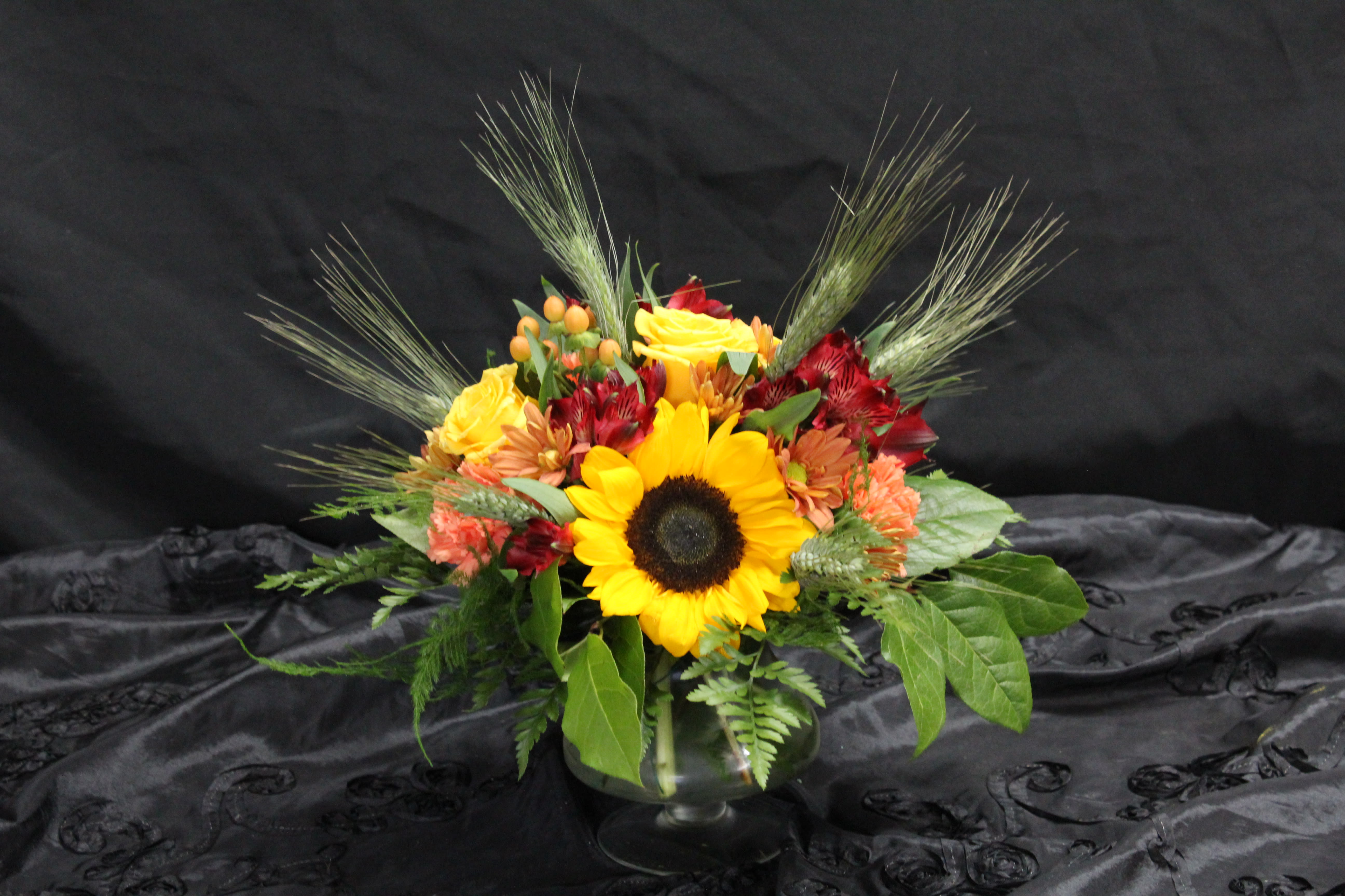 Autumn Wheat - Low grouping of sunflowers, roses, wheat, peruvian lilies,  hypericum berries, and carnations come together to create a festive floral arrangement.