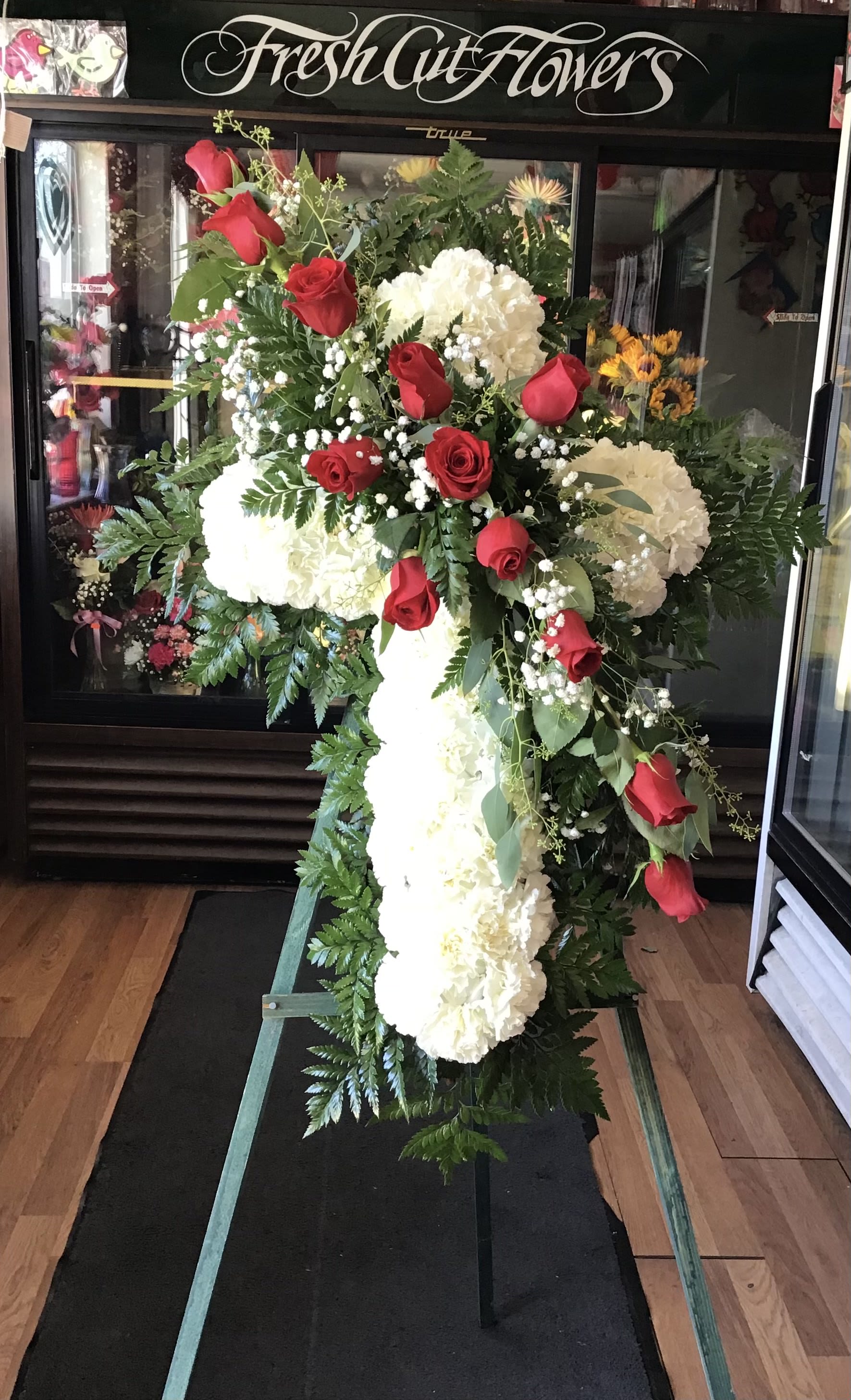 Faith &amp; Love Standing Cross - A classic sympathy Cross featuring fresh carnations accented with a beautiful line of roses cutting across the arrangement creating a graceful and heartfelt tribute.