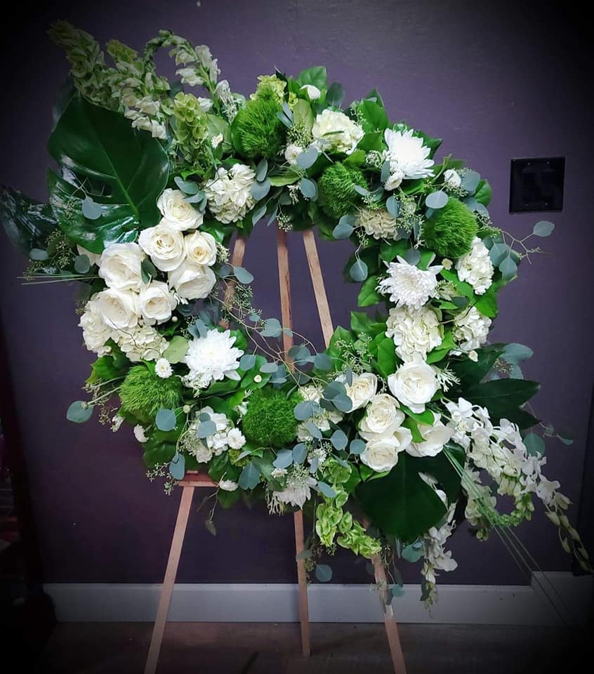 Large white and green standing wreath with roses, mums, orchids, and large glossy green leaves on a wooden easel.