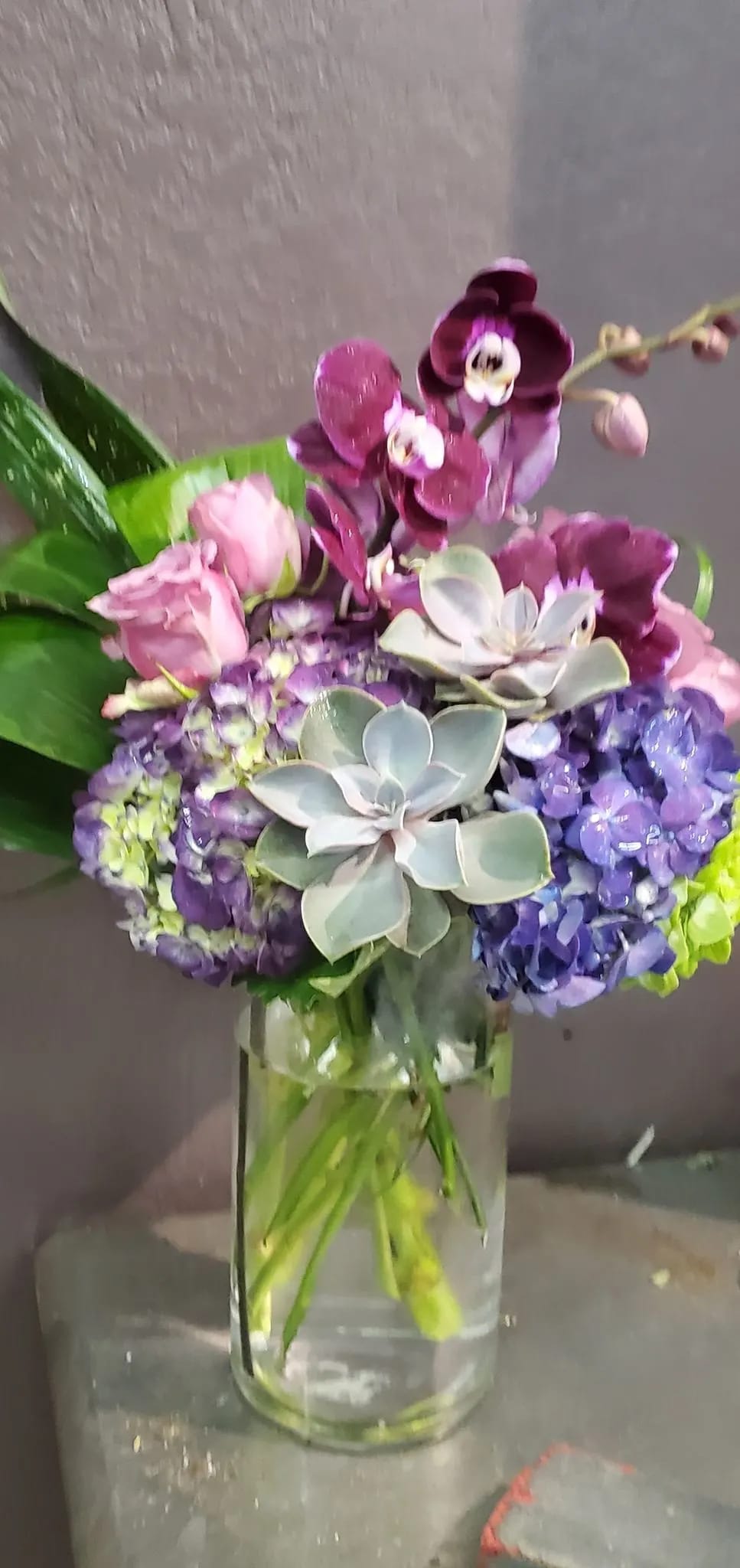 Tall glass vase arrangement with purple orchids, blue hydrangeas, pink roses, and succulents