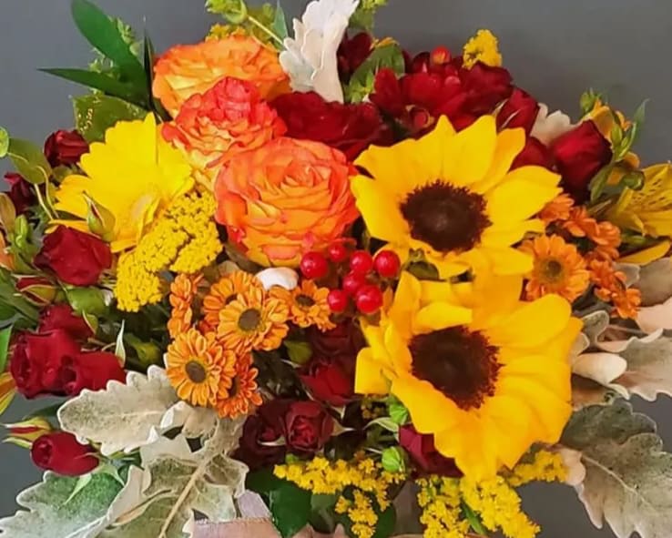 Bouquet of yellow sunflowers, orange and red roses, and orange mums with silvery foliage.