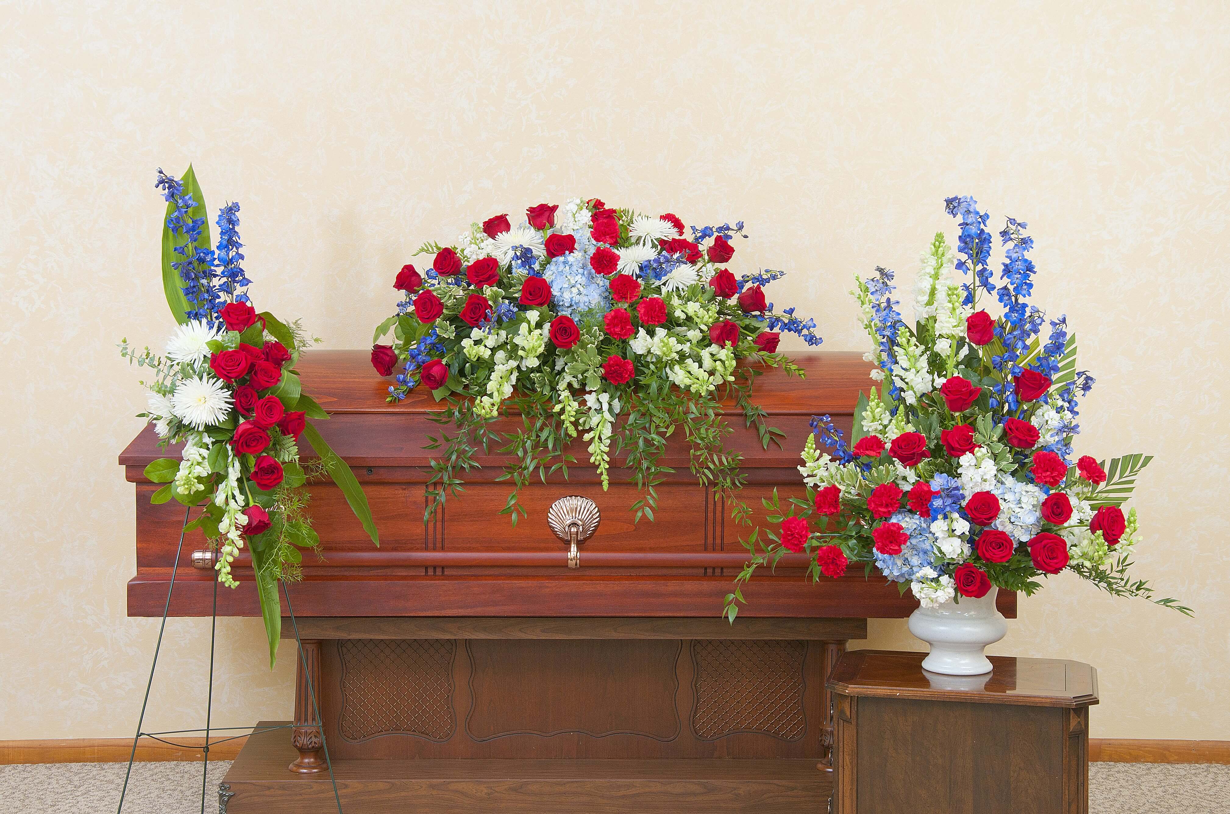 Red, white, and blue funeral floral set with casket spray and side arrangements on a wooden casket