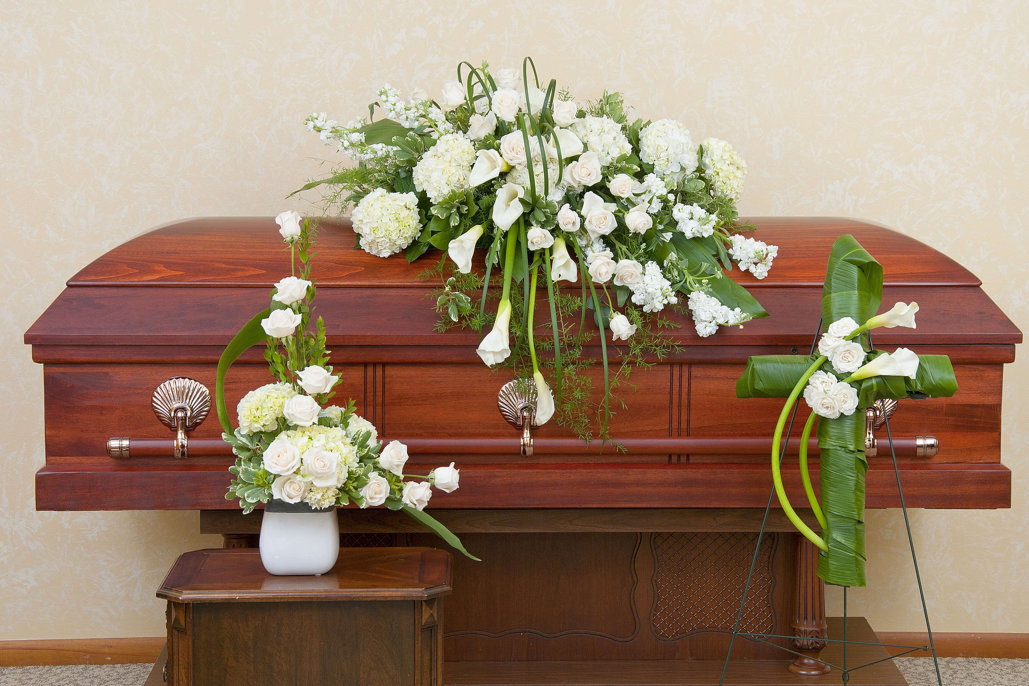 White funeral spray, vase bouquet, and cross arrangement displayed on a wooden casket