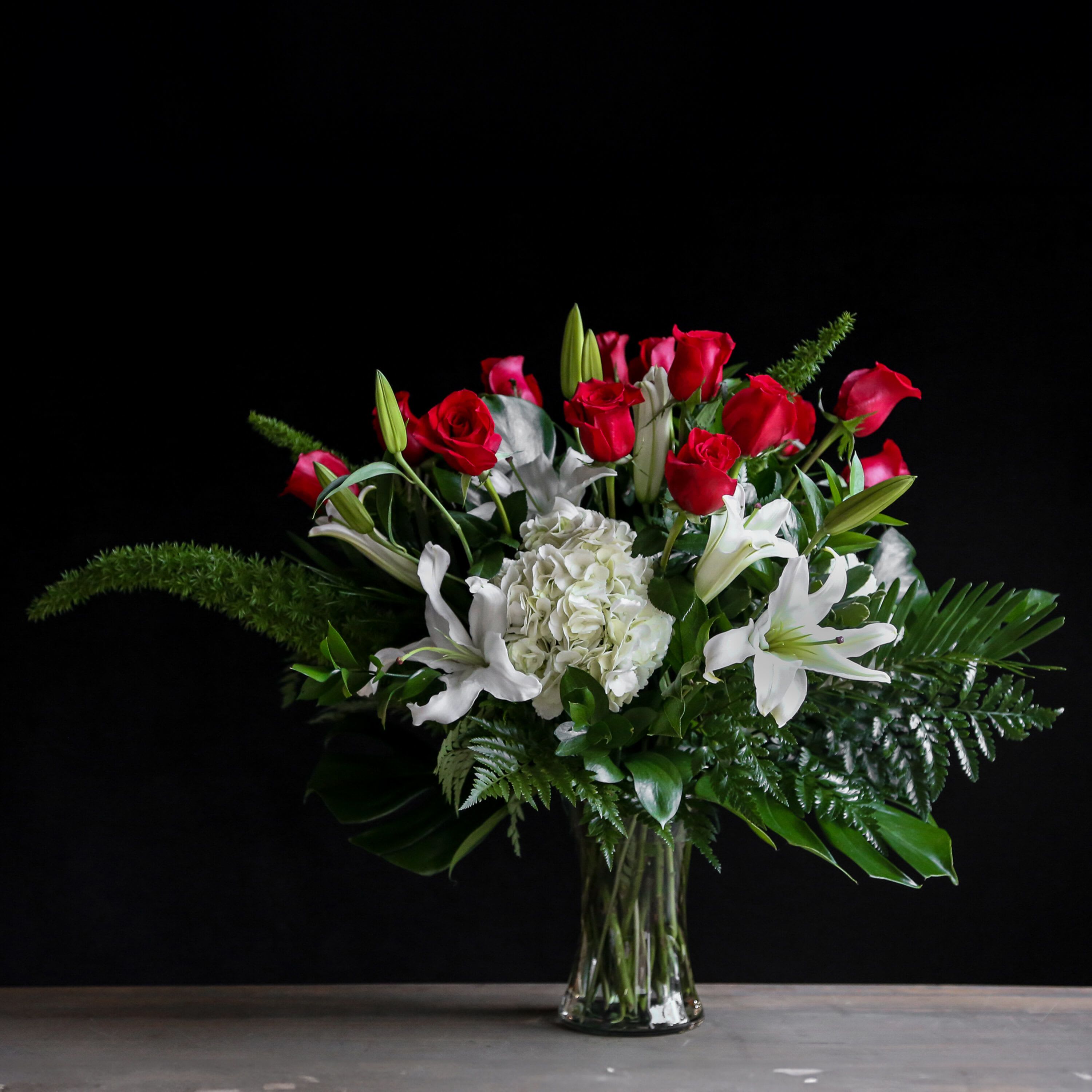 Tall arrangement of red roses, white lilies, and white hydrangea with arching green fronds in a clear vase.