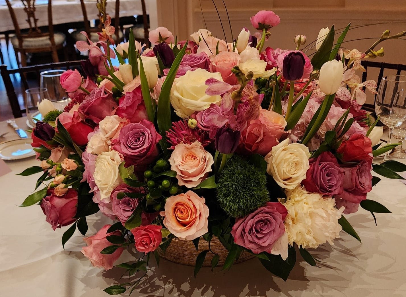 Low centerpiece of pink, purple, peach, and white roses with tulips and mixed blooms in a round bowl vase.