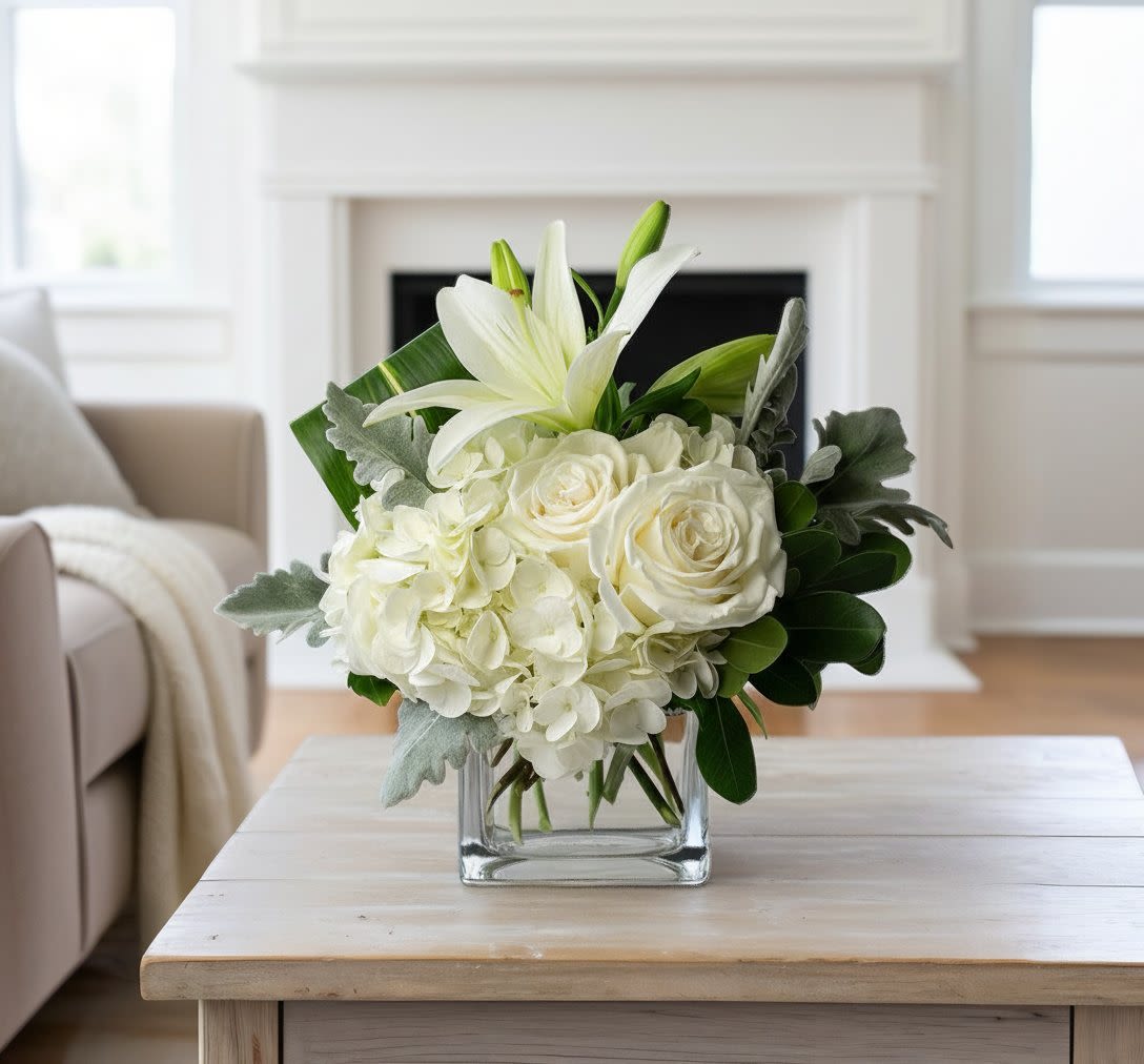 Low white arrangement of lilies, roses, and hydrangeas in a clear glass cube vase on a light wood table