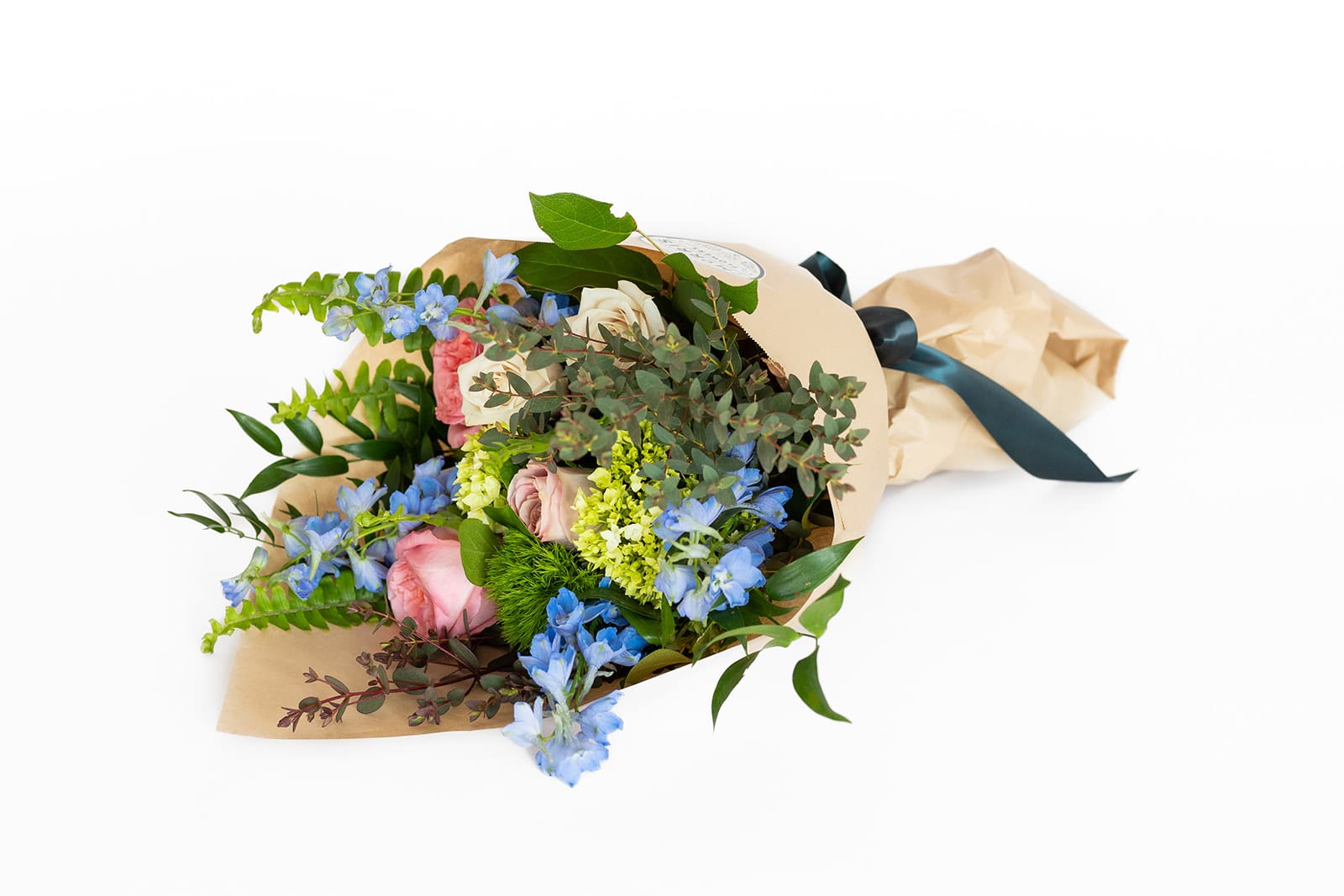 Wrapped bouquet of pink and cream roses with blue accent flowers in kraft paper tied with a dark ribbon