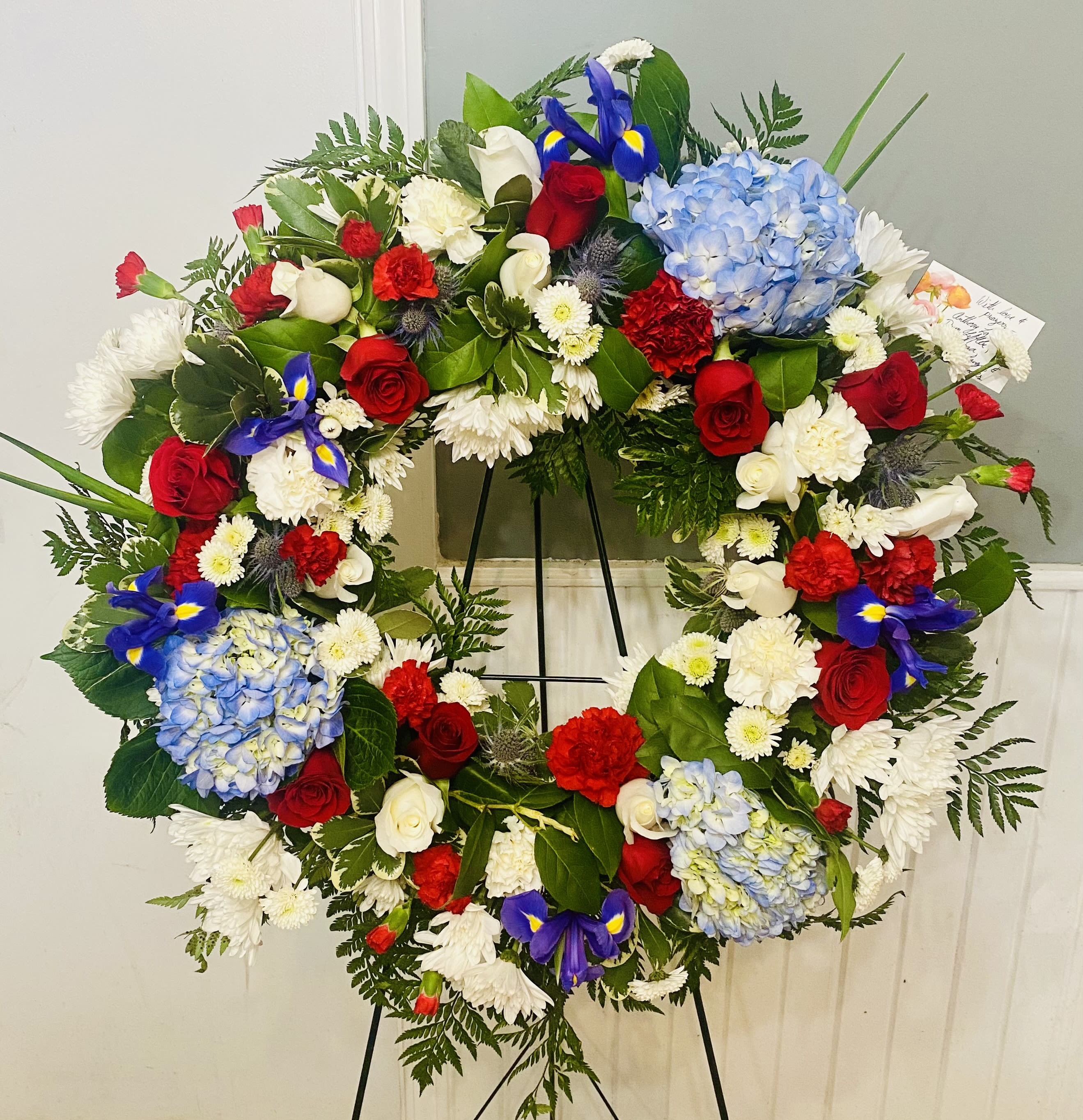 Red White and Blue Sympathy Wreath - A bold, patriotic sympathy wreath designed for memorial and ceremonial tribute, this red, white and blue funeral wreath expresses solemn honor with visual clarity. The circular arrangement and the inclusion of a metal easel stand make it an appropriate sympathy arrangement for a graveside service, memorial ceremony, or funeral home display-an elegant way to show respect and remember their service or spirit.  Tightly arranged red roses and red carnations contrast with crisp white roses and white chrysanthemums, while pale blue hydrangeas and blue iris add cool highlights across a dense bed of deep green foliage. Small daisy-like blooms fill the surface for texture, creating a layered, formal presentation that reads clearly from a distance. The wreath arrives ready to stand on the included black metal easel and comes with a card for your personal message.  Made at a substantial scale to suit public memorials as well as private services, this arrangement is crafted to maintain its shape and color impact throughout viewing. It's a meaningful way to honor someone who cherished patriotic colors or to mark occasions like Veterans Day, Memorial Day, or a funeral-offer a timeless, respectful tribute that lets the person you're remembering feel truly honored.