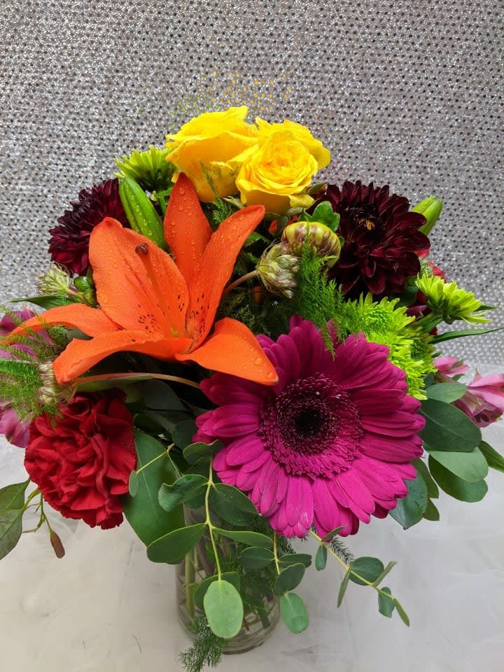 Colorful bouquet with orange lily, pink gerbera, yellow roses, and red carnations in a glass vase