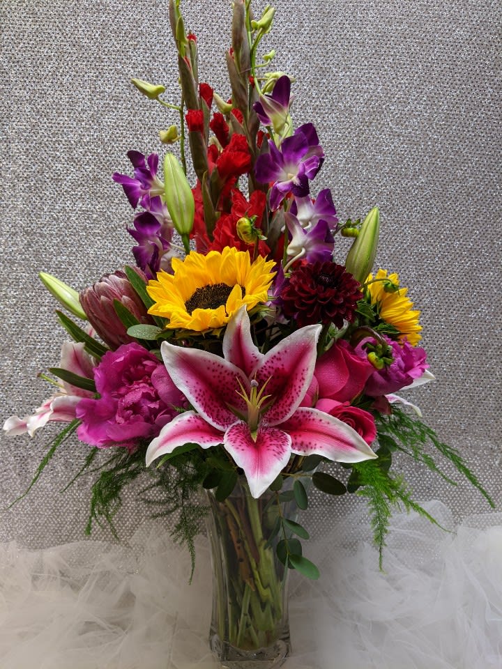 Tall mixed bouquet with pink lilies, sunflowers, purple orchids, and red blooms in a clear glass vase