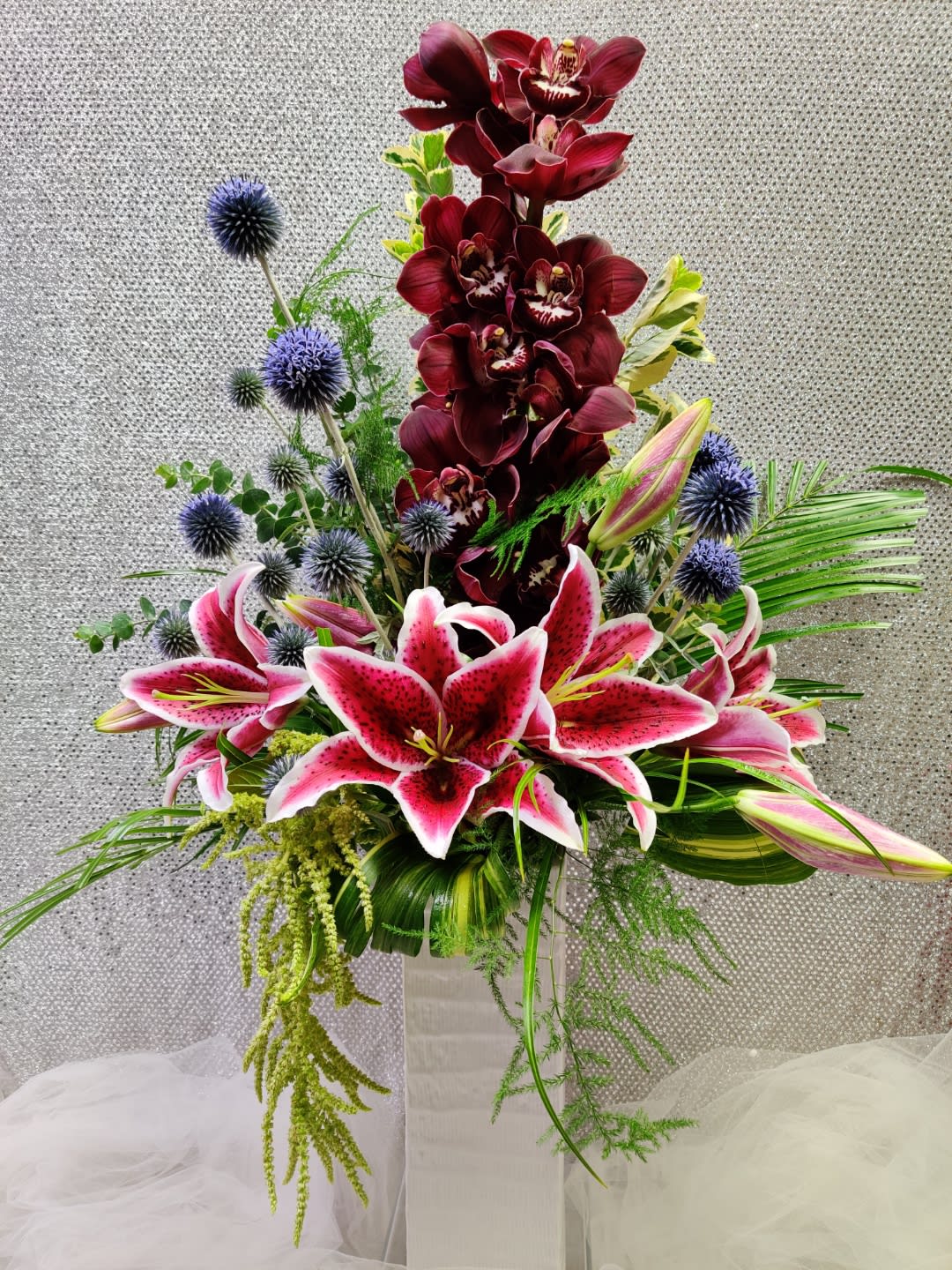 Tall floral arrangement with pink lilies, burgundy orchids, and blue thistle in a vase