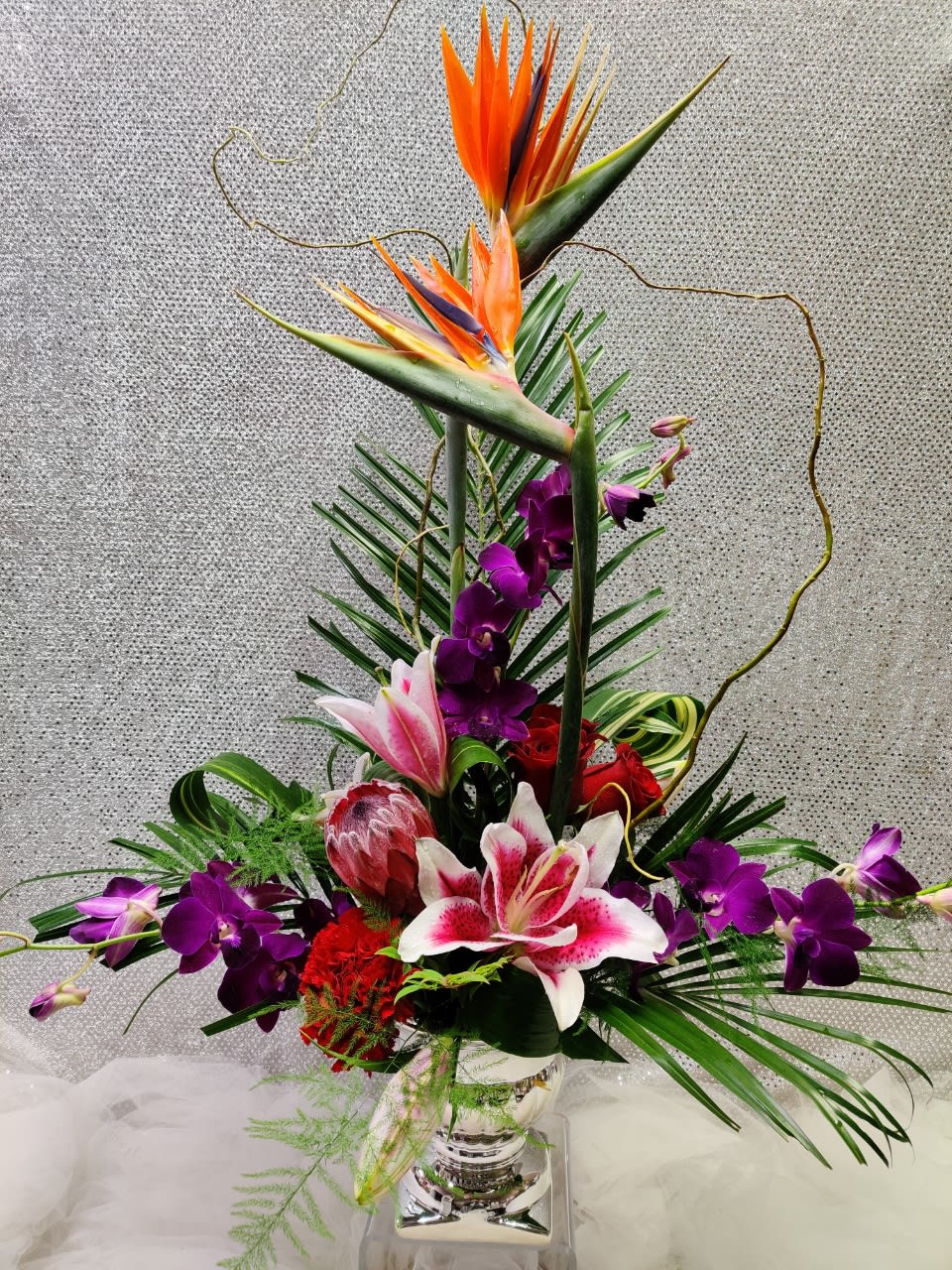 Tall tropical flower arrangement with orange bird of paradise and purple orchids in a glass vase