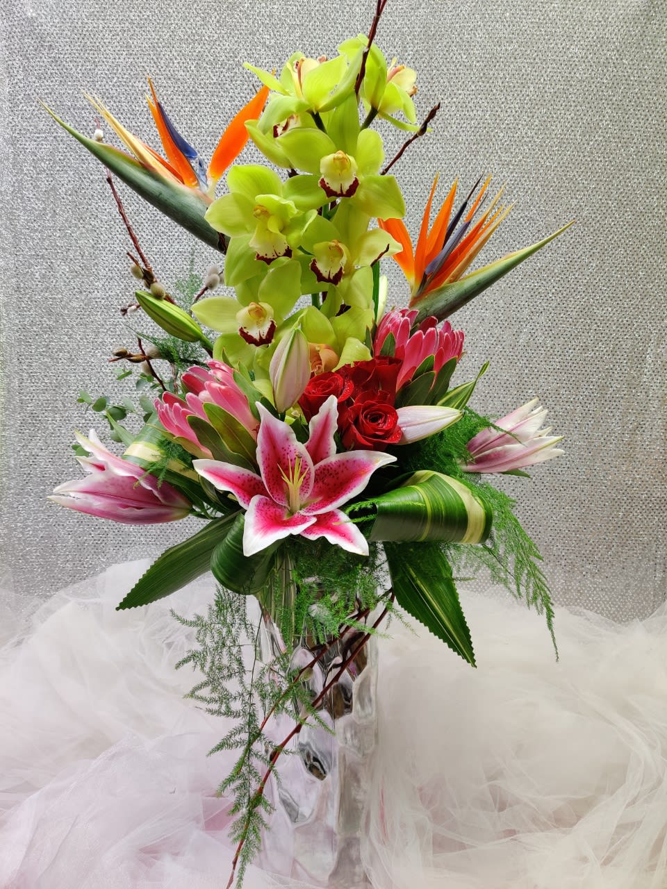 Pacific Paradise - Includes Cymbidium, Birds of paradise, Stargazer lilies, Protea, Roses, pussy willow and Tropical Greenery in a tall Silver Vase.