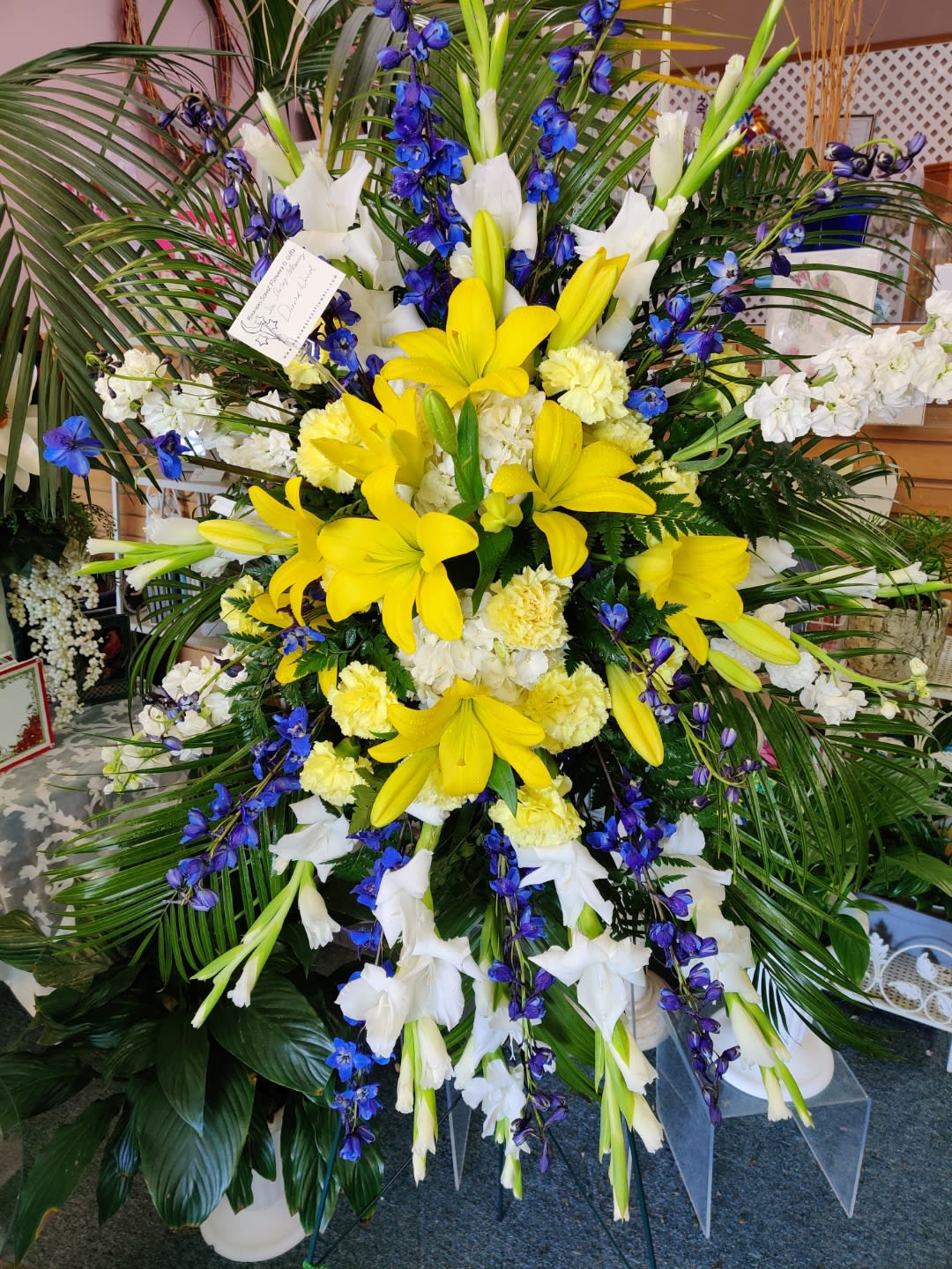 Large standing floral spray with yellow lilies, white blooms, and blue flowers