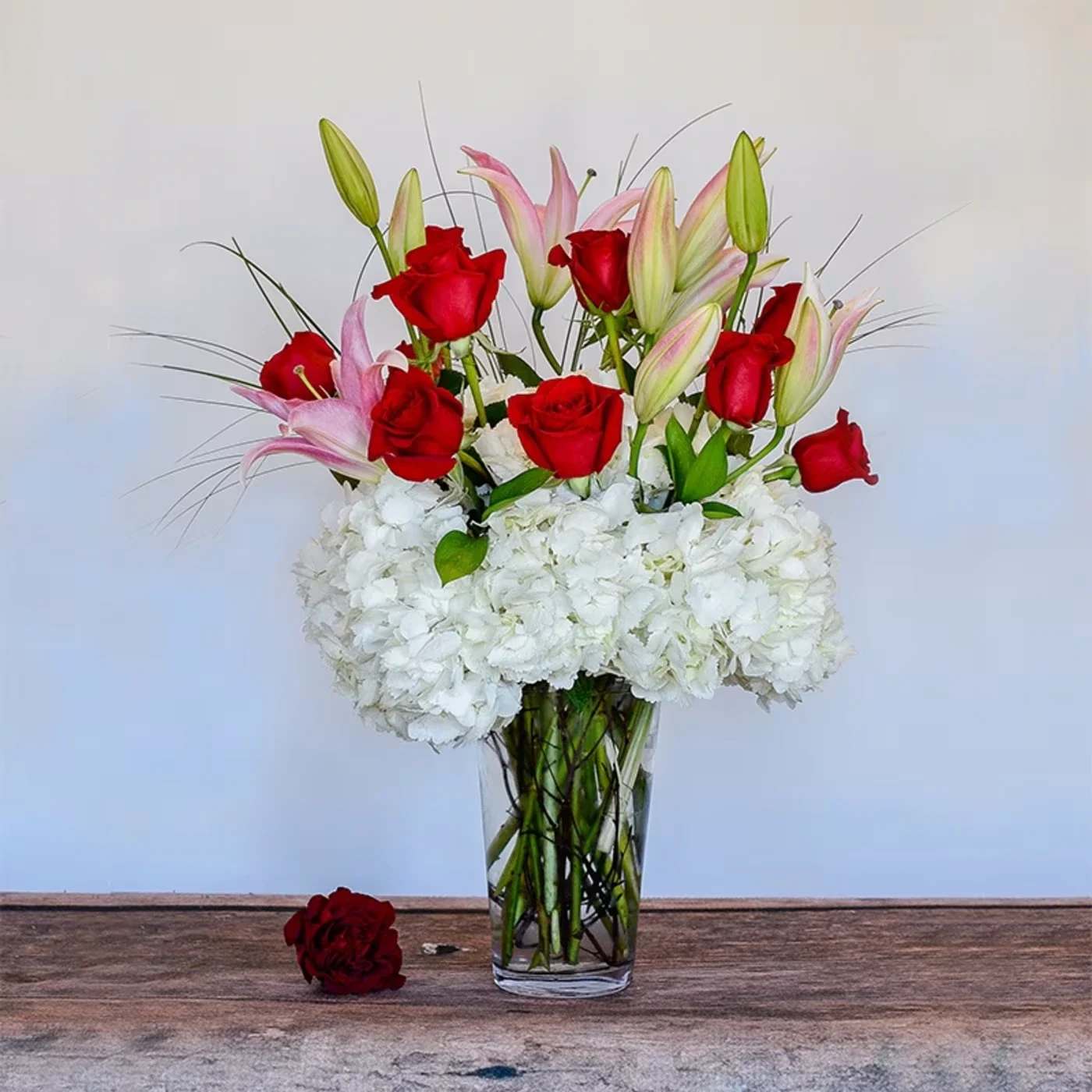Tall vase arrangement of red roses, pink lilies, and white hydrangeas with one loose rose beside it.