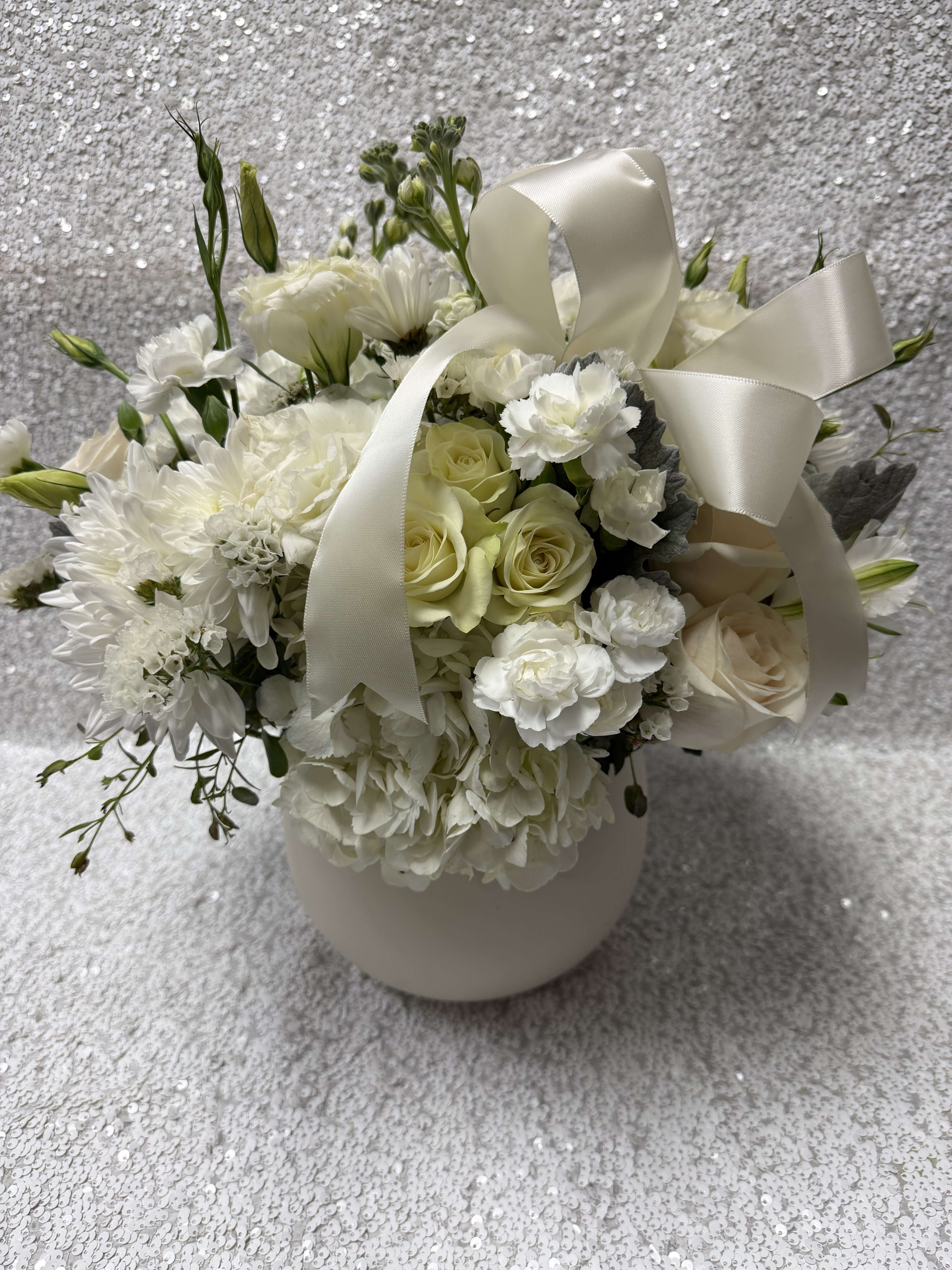 White floral arrangement with roses, carnations, and a satin ribbon in a vase