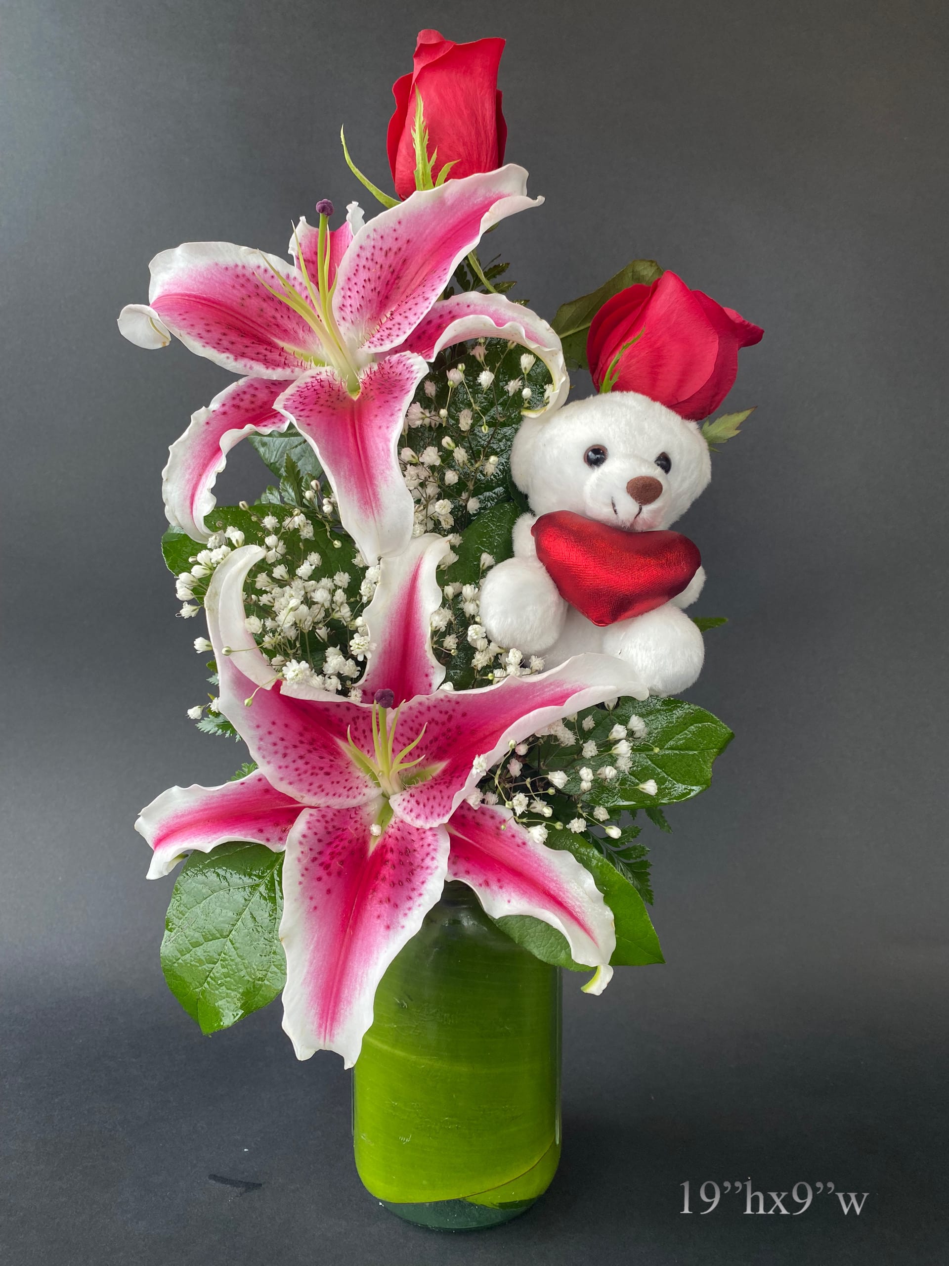 Pink lilies and red roses in a green vase with a teddy bear holding a heart