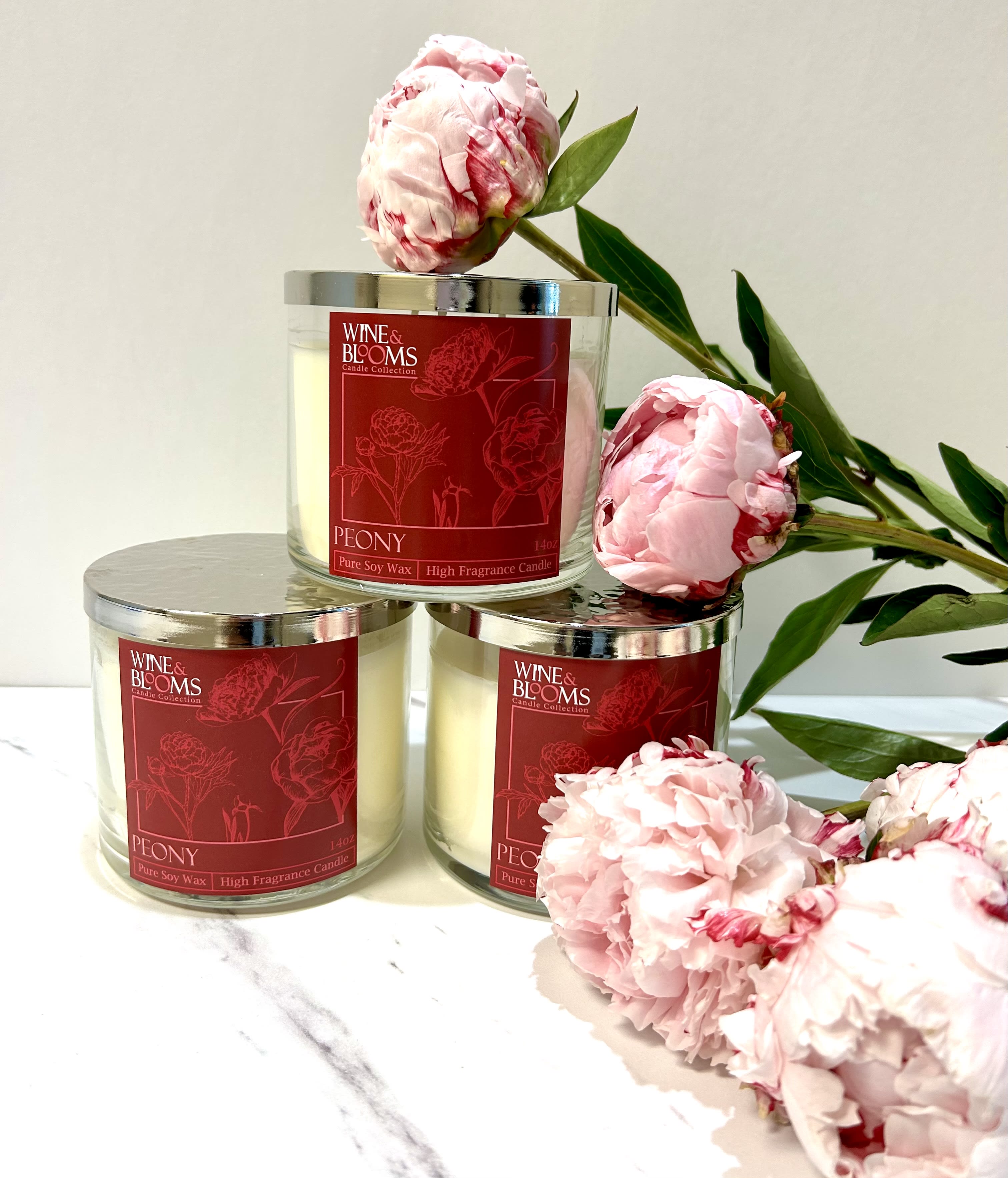 Three peony-scented candles with pink peony blooms arranged around them