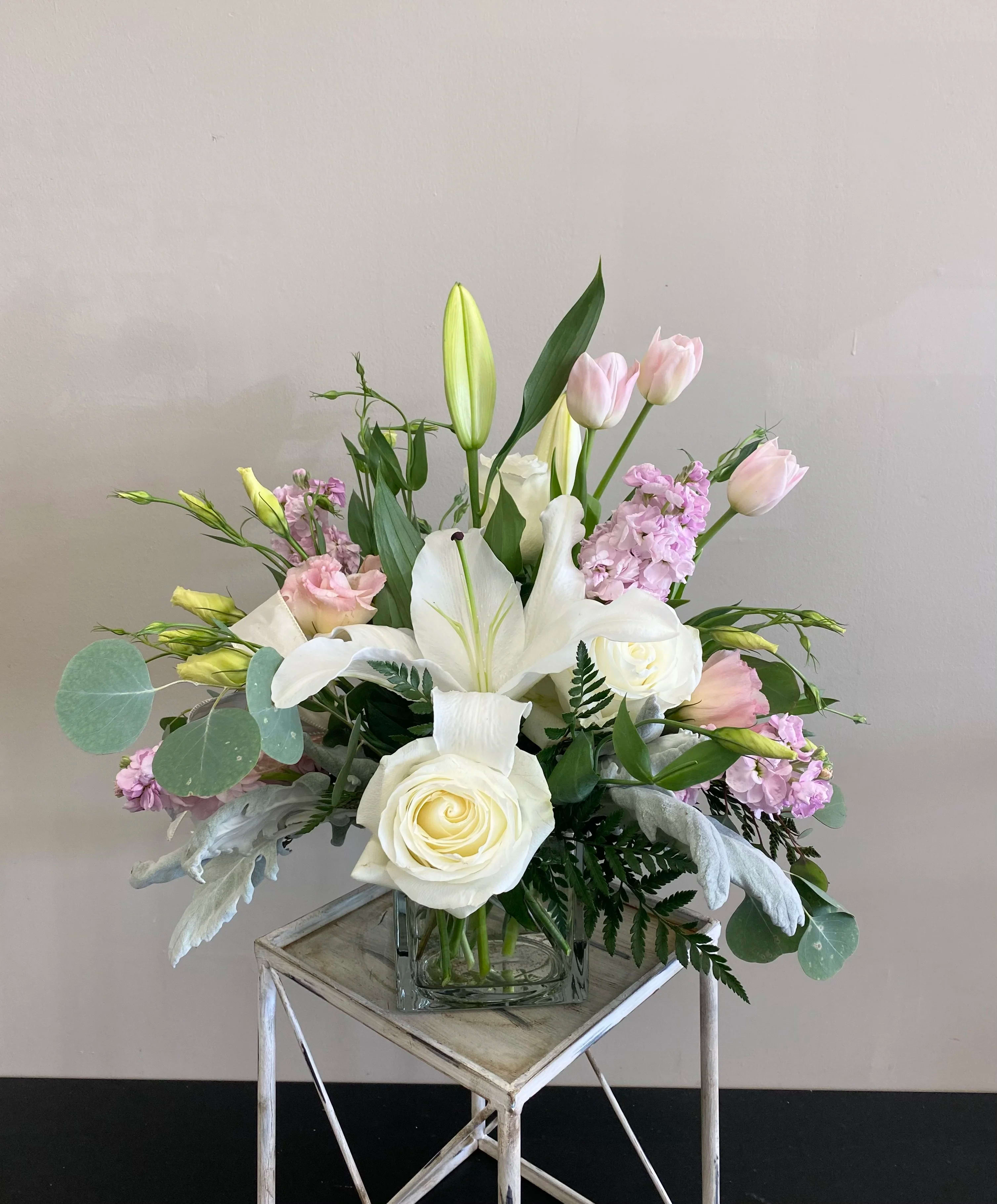 Pastel arrangement of white lilies and roses with pink tulips and stock in a glass cube vase.