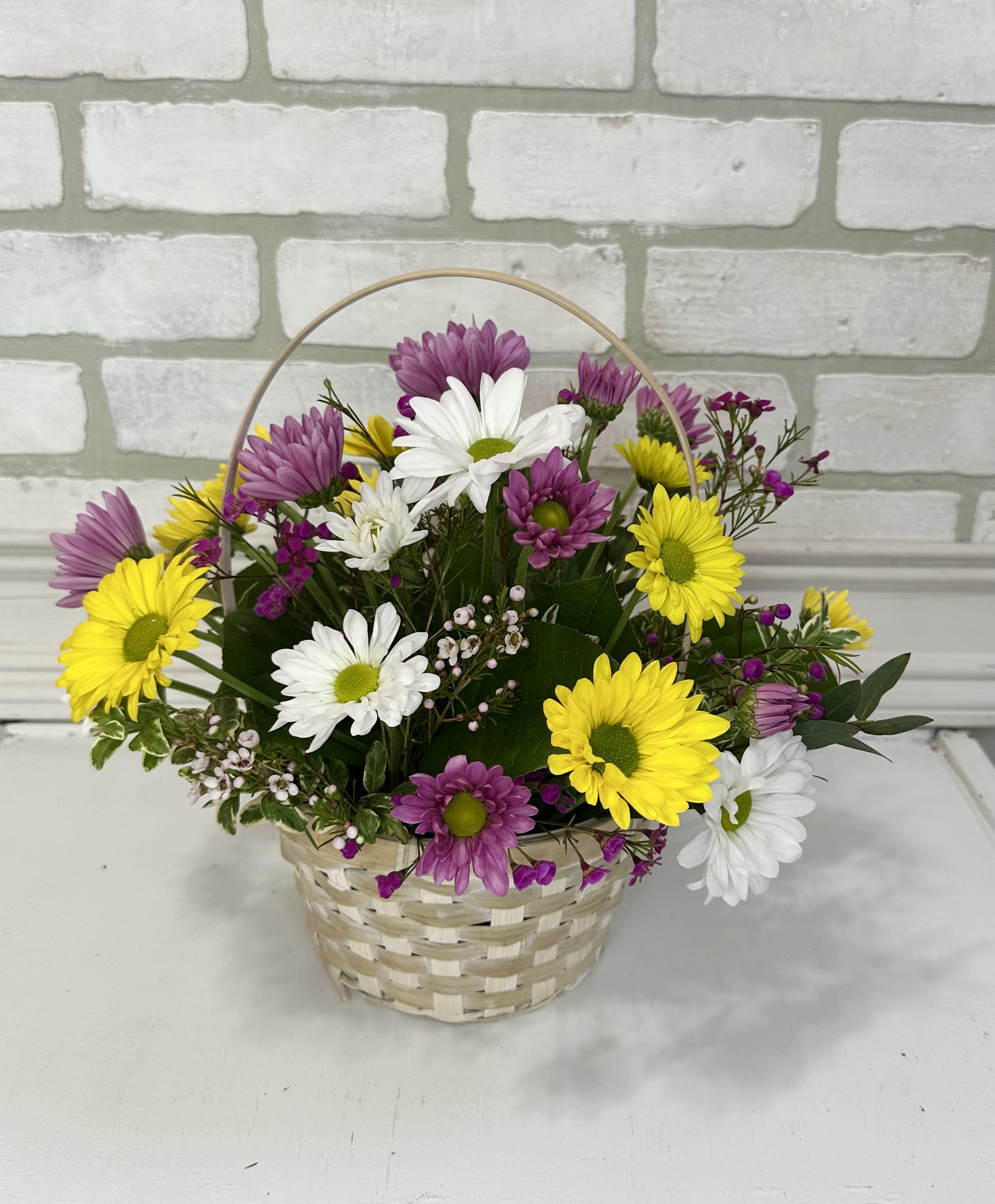 Basket arrangement of yellow, white, and purple daisies with small pink filler flowers.