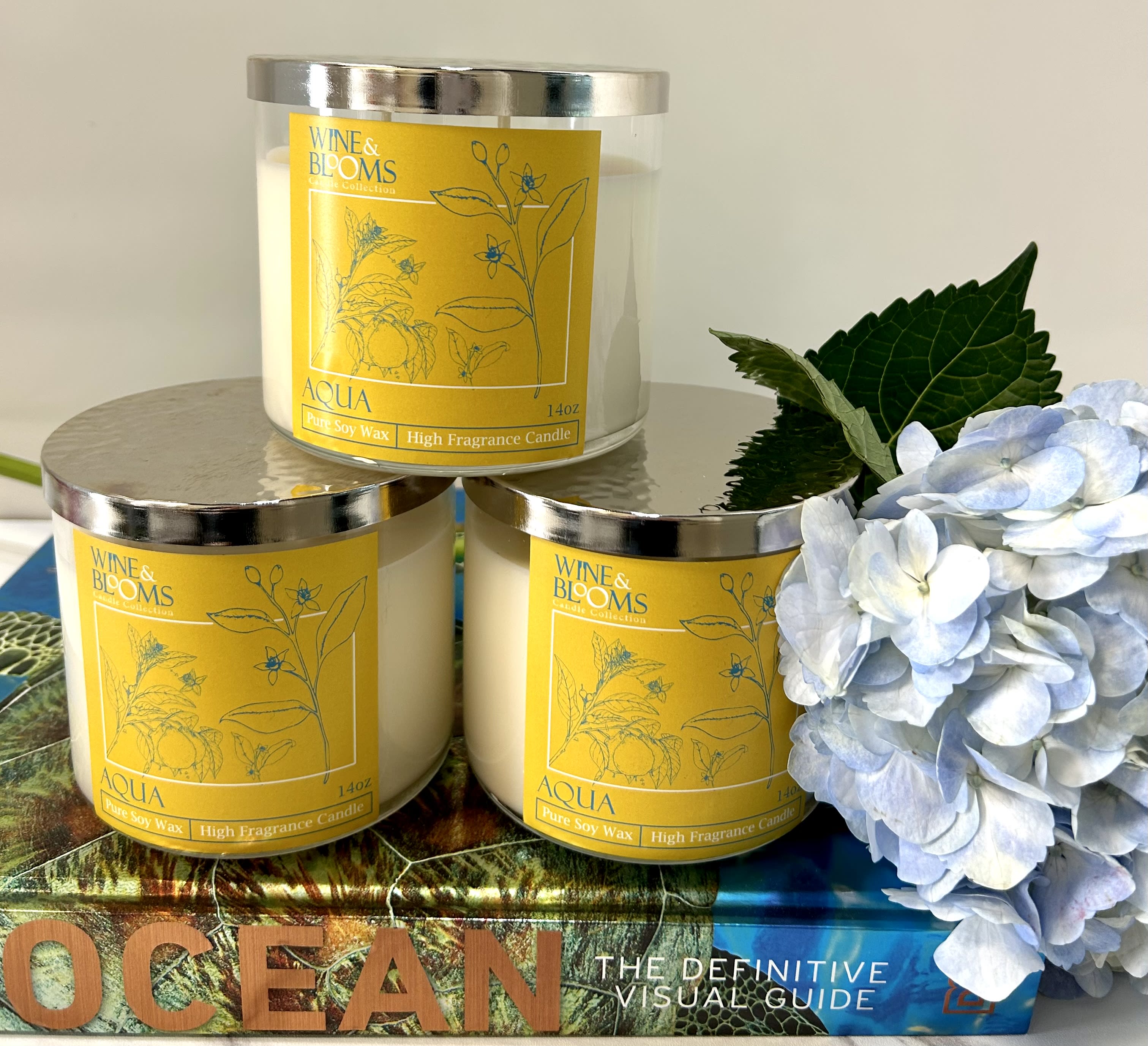 Three scented candles beside pale blue hydrangeas and a book