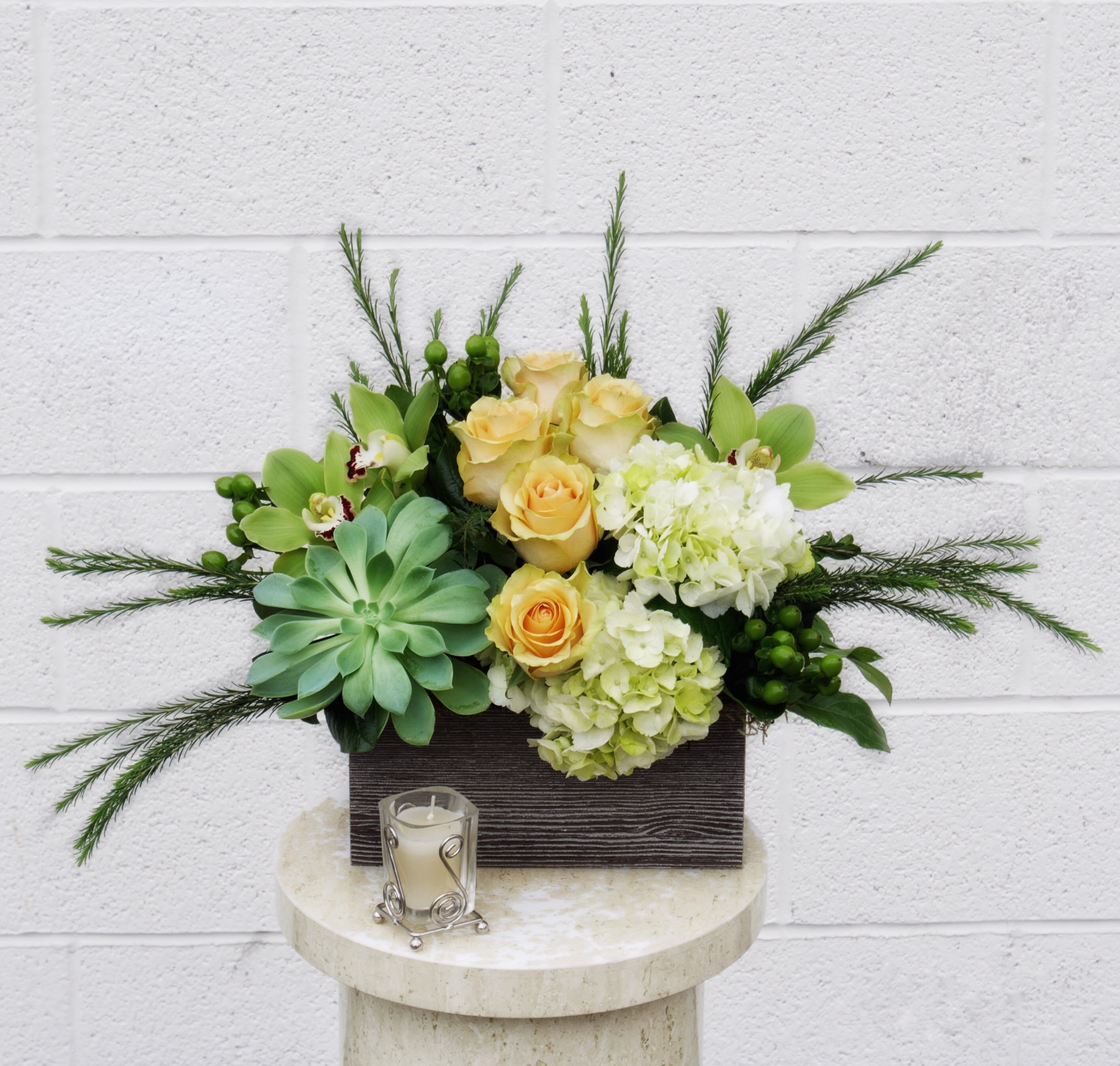 Arrangement of yellow roses, hydrangeas, orchids, and succulents in a rectangular container with a candle