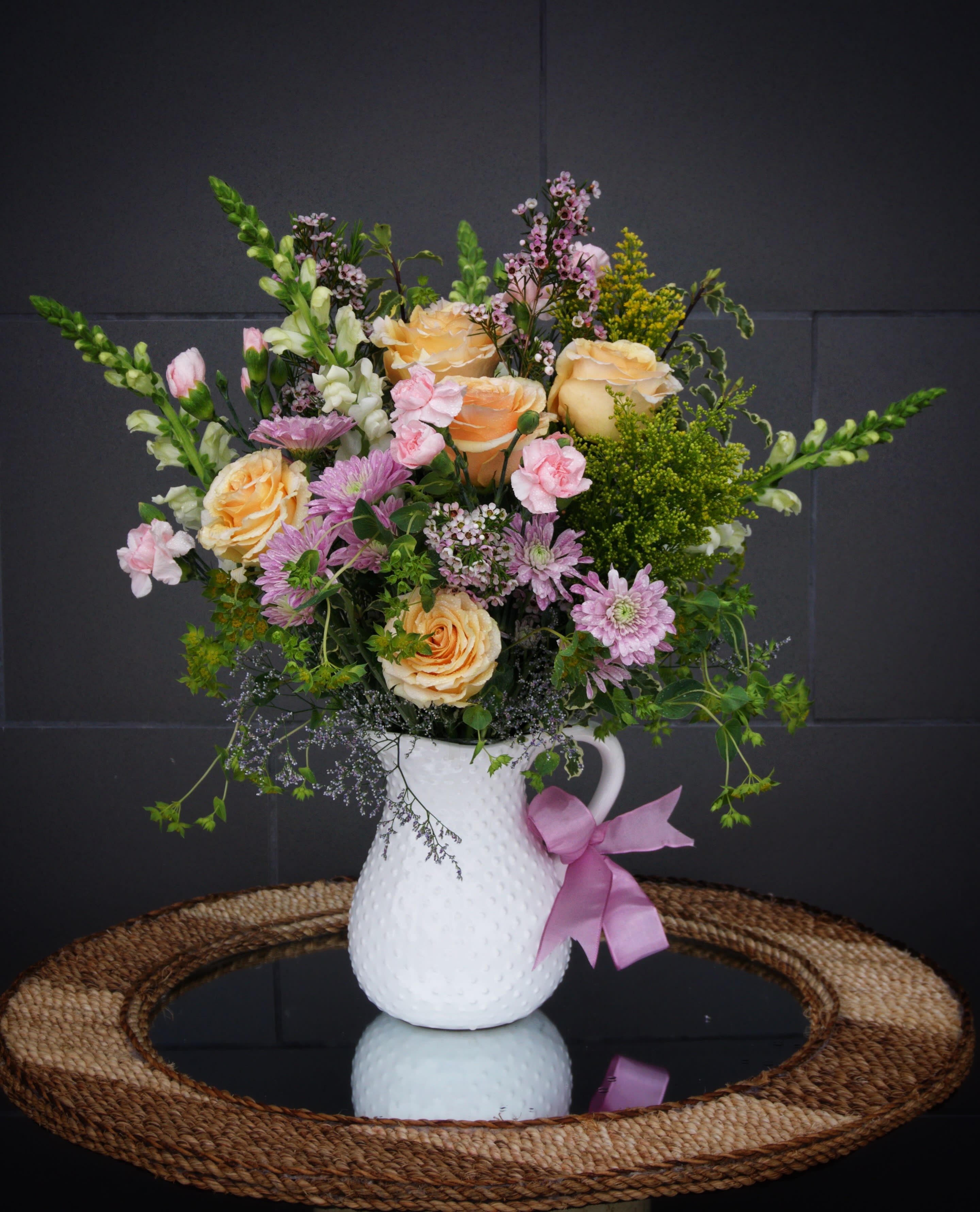 Peach roses and pink flowers in a white pitcher vase with a pink ribbon