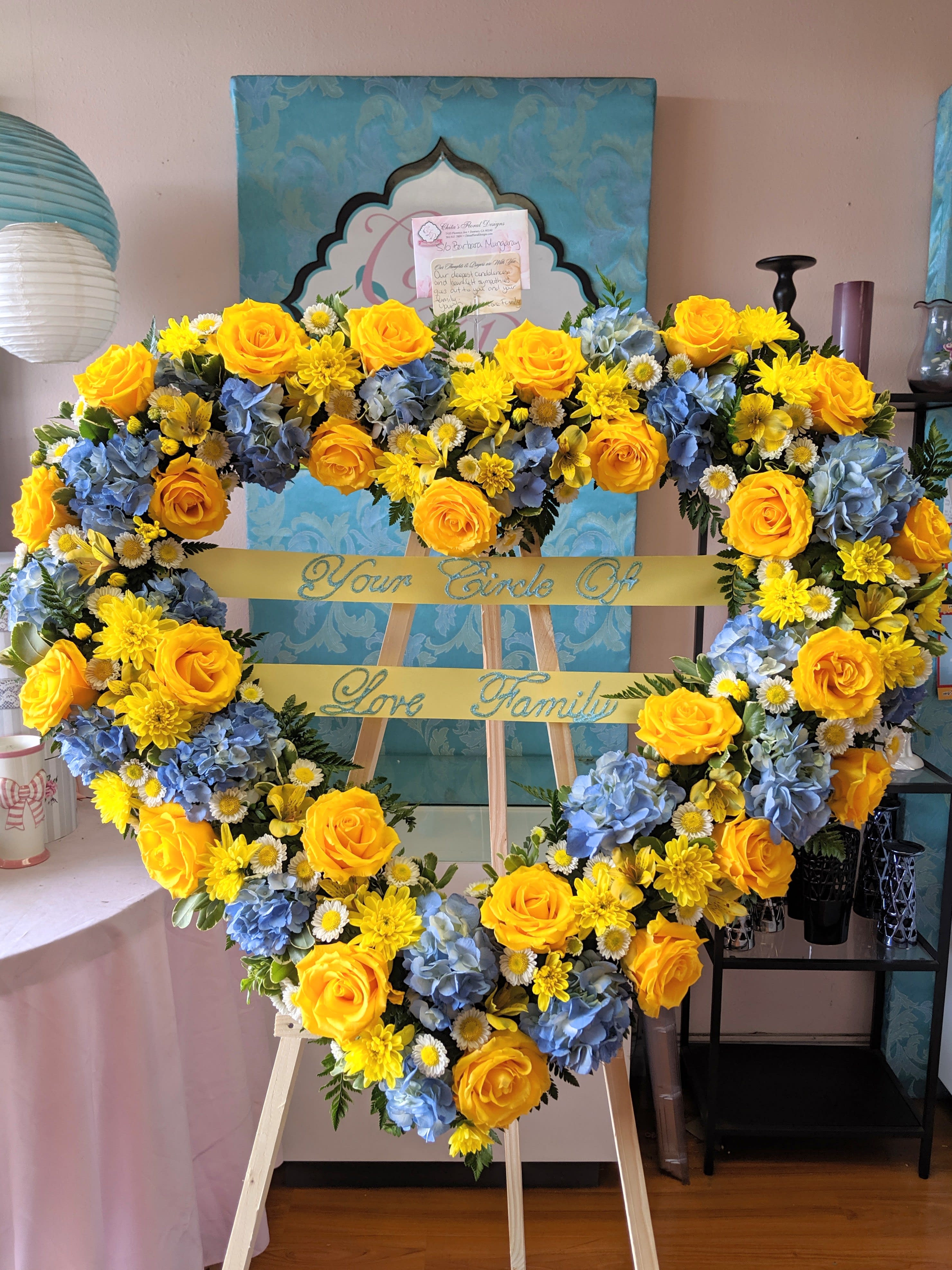 Heart-shaped floral wreath with yellow roses, blue hydrangeas, and daisies on an easel