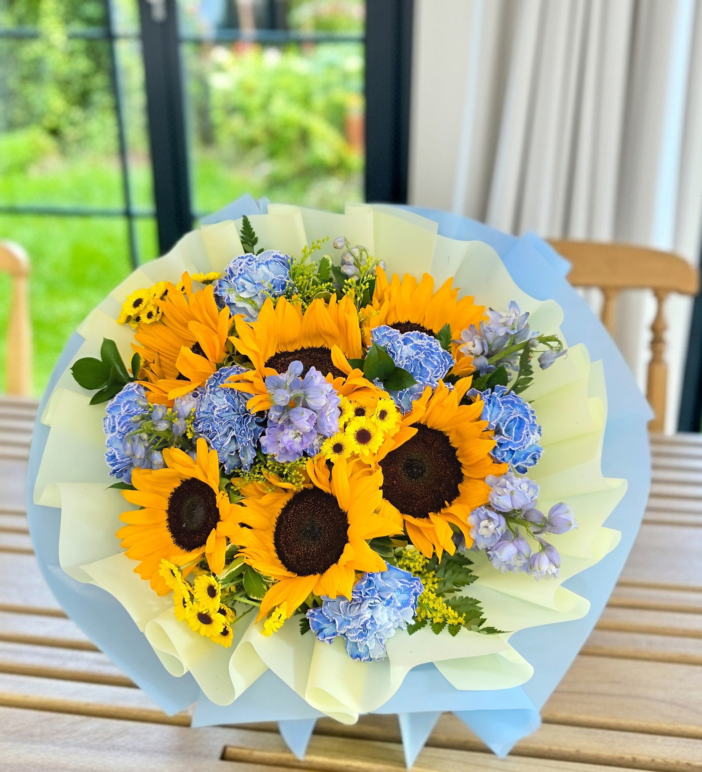 Sunshine Wrapped Bouquet  - Bright sunflowers take center stage in this Sunny Days bouquet, a cheerful flower arrangement perfect for birthdays, congratulations, or simply letting someone know you're thinking of them. The combination of bold yellow blooms with clusters of blue hydrangeas and touches of purple and white creates an uplifting, celebratory look that makes the recipient feel genuinely seen.