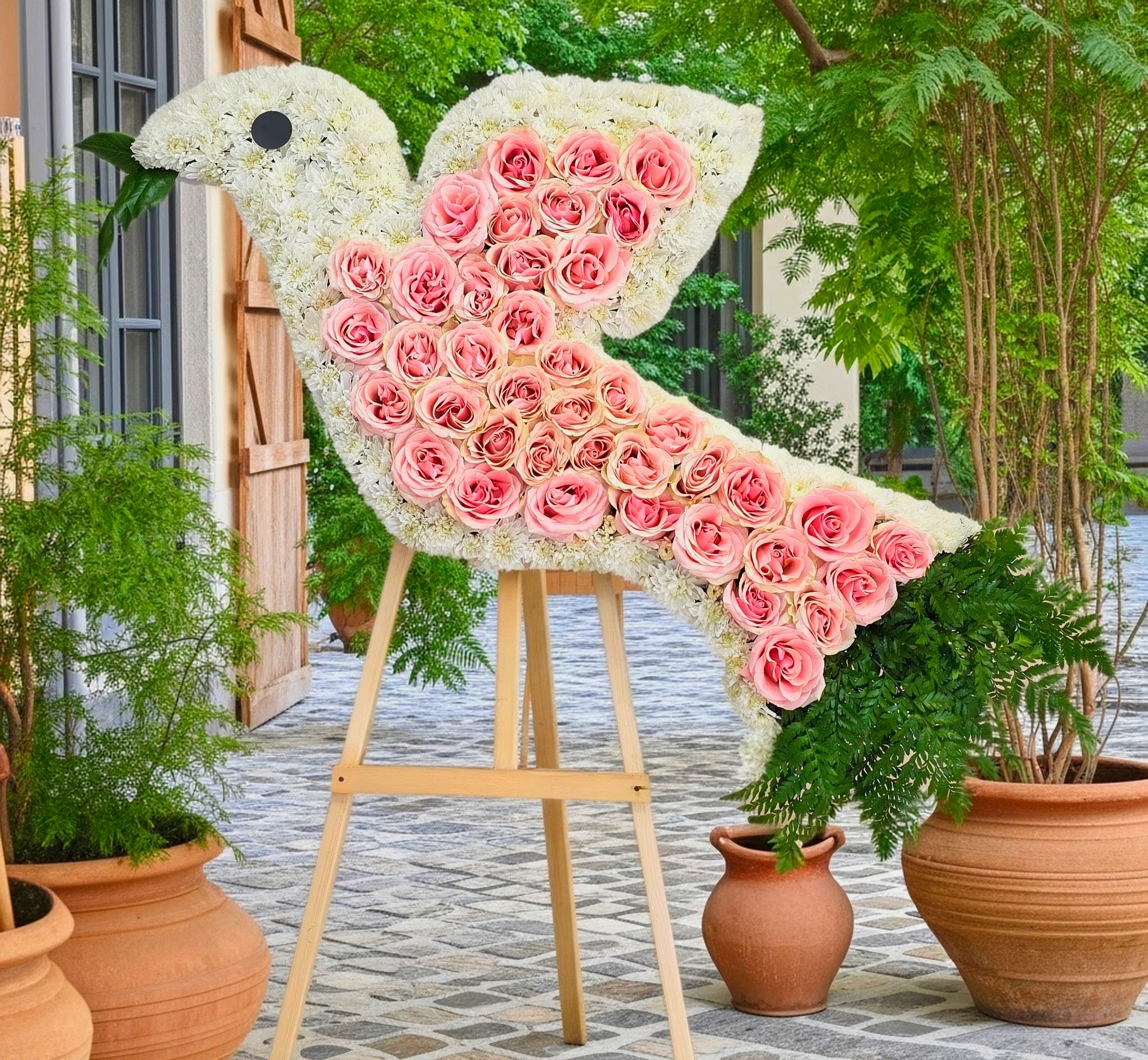 Peaceful Dove Standig Sympathy - A sculptural dove-shaped standing spray that reads soft, serene, and quietly celebratory-perfect as a sympathy arrangement or memorial tribute. The Peaceful Dove standing spray uses a white floral body to convey calm and comfort, while a wing and tail of pink roses introduce a gentle, romantic accent that honors the person you're remembering. The playful black eye detail and graceful bird silhouette make this arrangement feel intentional and comforting. Handcrafted on a wooden easel, the spray combines dense white blooms with clusters of pink roses and touches of green foliage for depth and texture; terracotta pots anchor the design and give it a grounded, natural presence suited to outdoor services or ceremony settings.