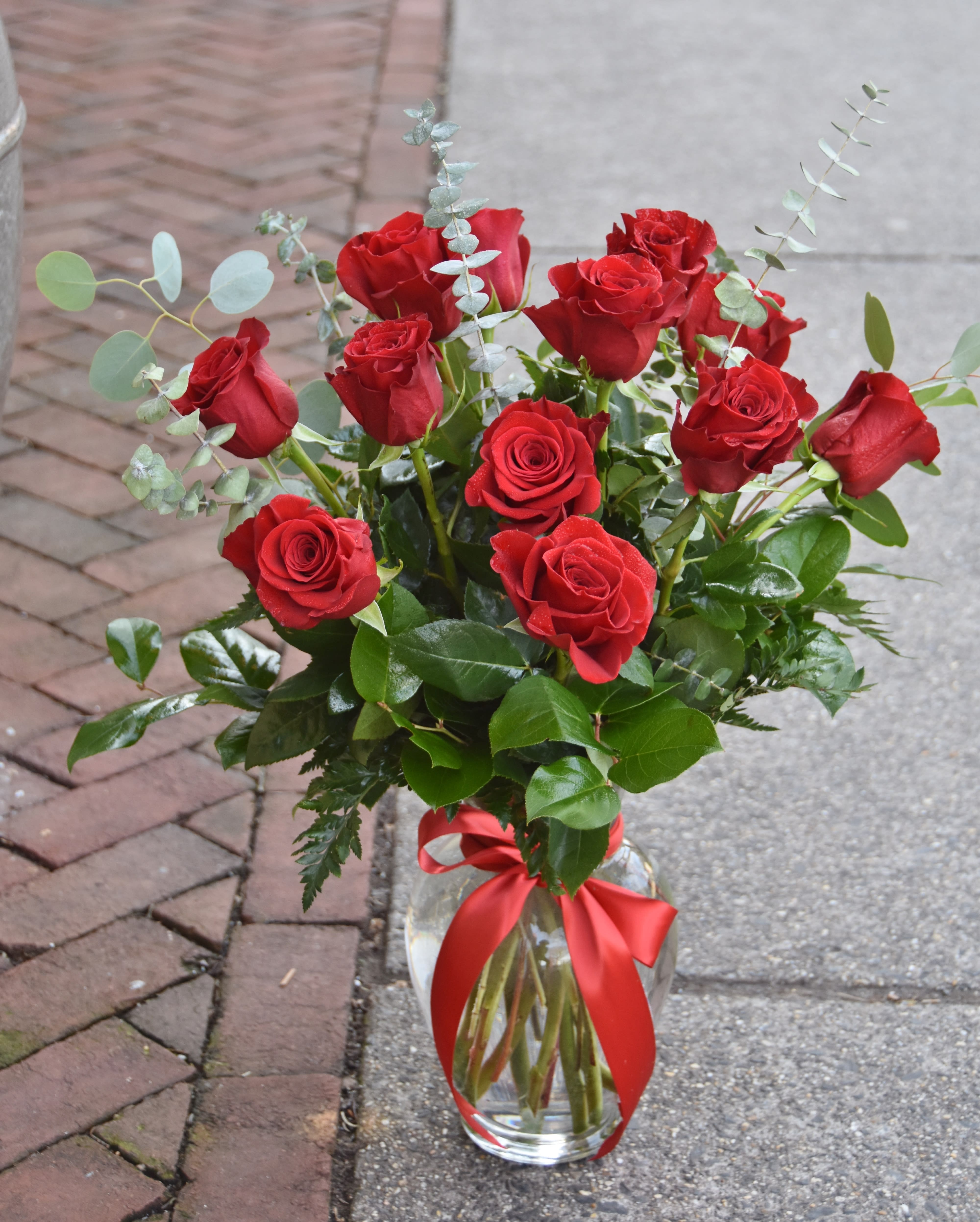 Arrangement of red roses with eucalyptus in a clear glass vase tied with a red ribbon.