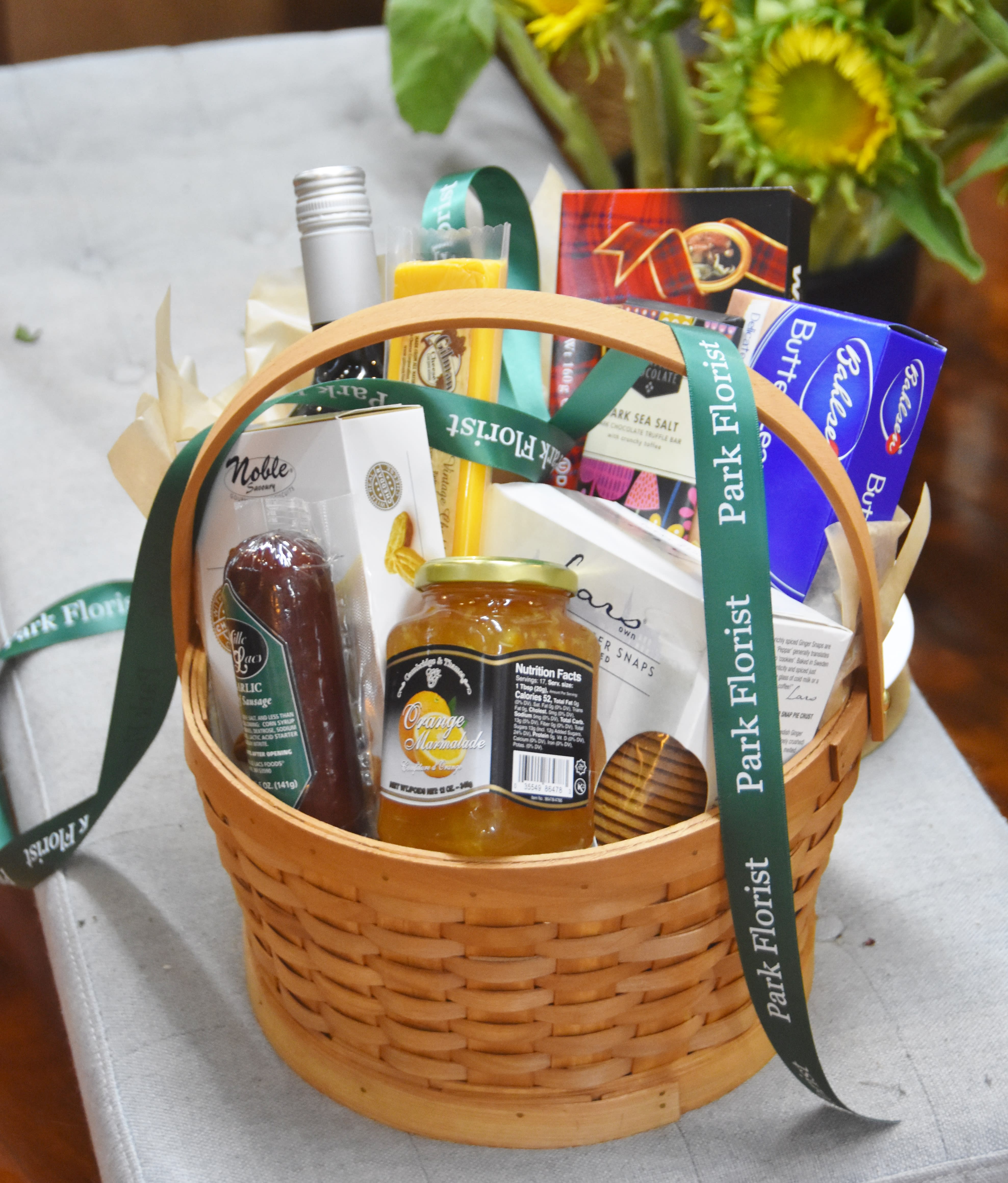 Gourmet food gift basket with marmalade, sausage, snacks, and a ribbon, with sunflowers in the background