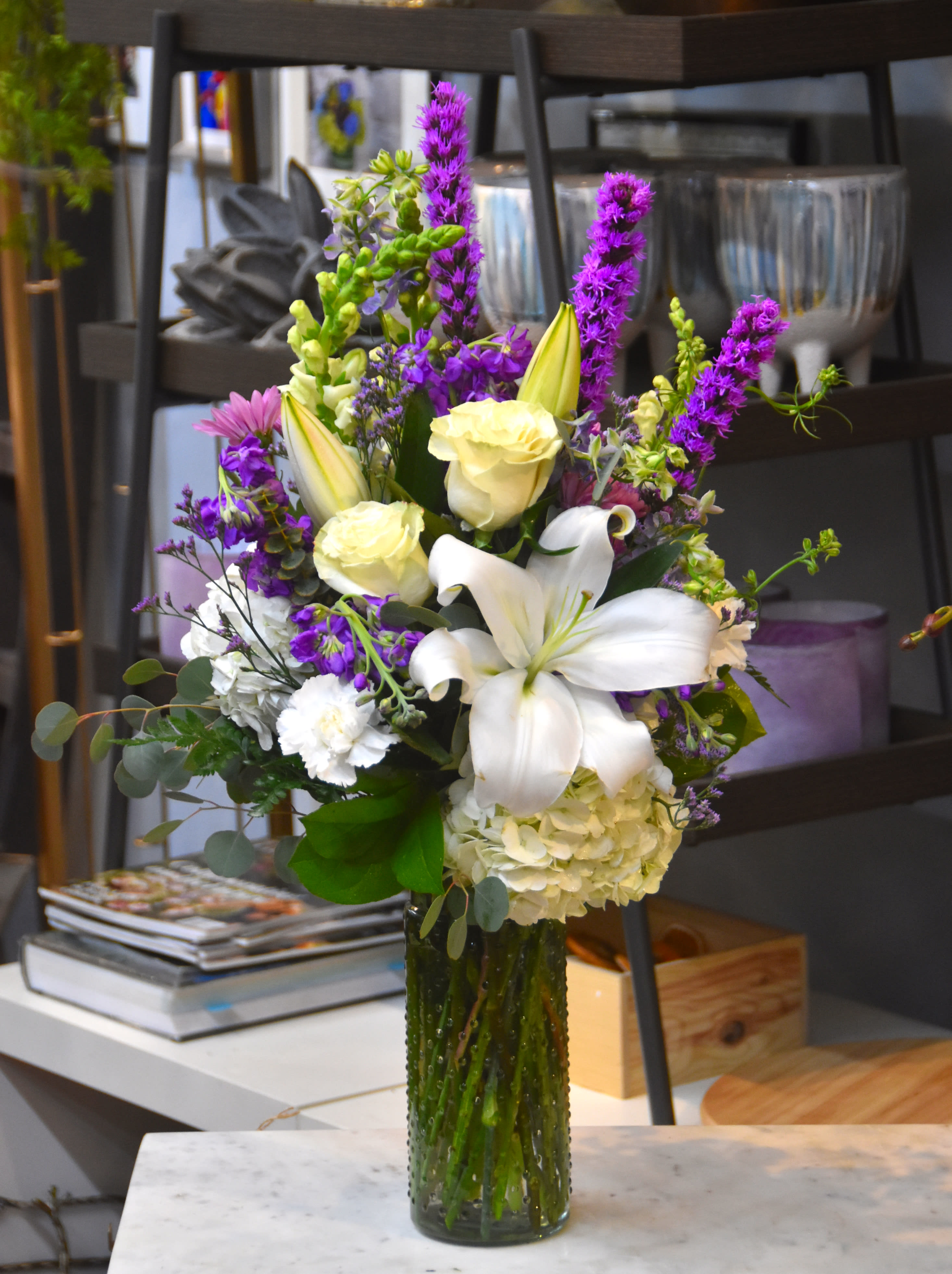 Tall arrangement of white lilies and roses with purple flowers in a green glass vase