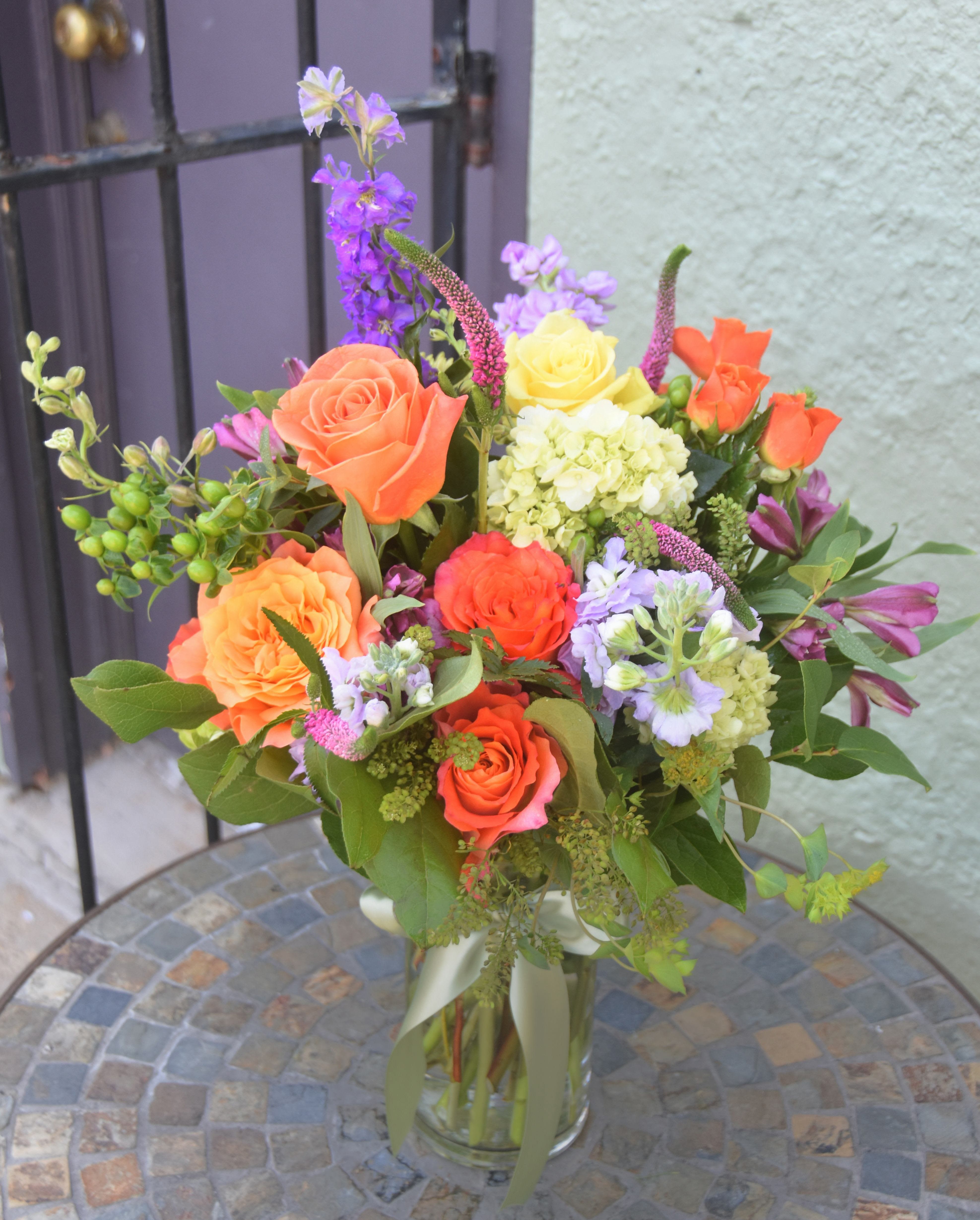Mixed bouquet of orange roses, yellow blooms, and purple flowers in a clear glass vase