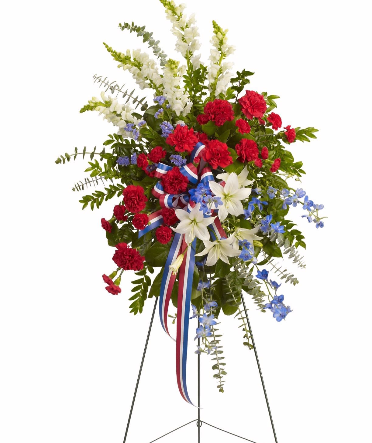 Patriotic standing spray with red carnations, white lilies and blue flowers on an easel with a tricolor ribbon.