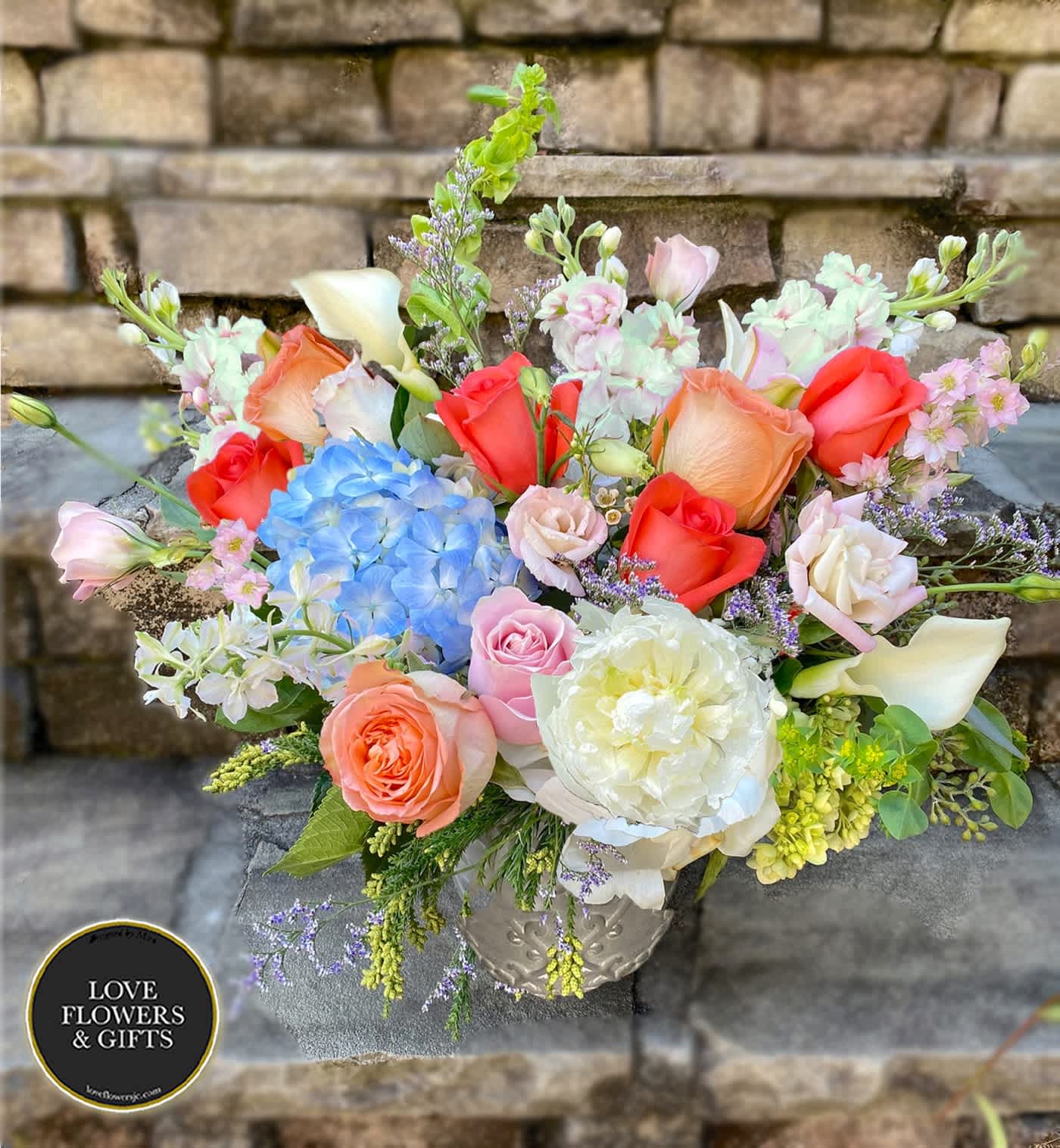 Mixed bouquet of peach roses, blue hydrangea, white peony and calla lilies in a patterned vase.