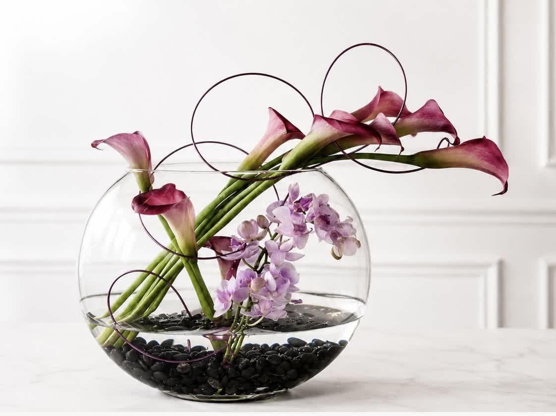Calla lilies and purple orchids in a round glass bowl with black stones