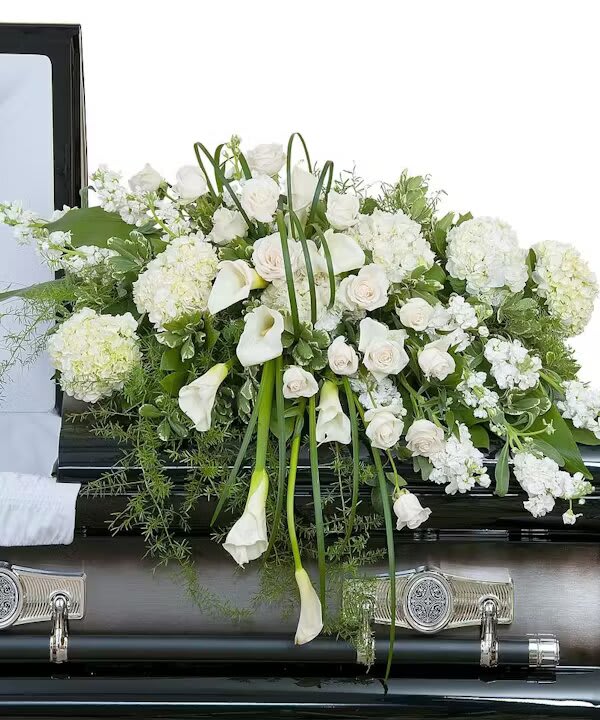 ELEGANT LOVE CASKET SPRAY - Elegant white premium flowers in a lush casket spray.  The Elegant Love Casket Spray is a testament to timeless beauty and grace, designed to convey a sense of peace and eternal love. This lush casket spray is meticulously composed of premium white flowers, each bloom carefully chosen for its purity and sophistication. In life's final farewell, this floral arrangement speaks volumes where words fall short, offering a silent yet profound tribute to the one who has passed.  Centered around the dignified beauty of white roses, which have long been associated with reverence and remembrance, the spray radiates a comforting glow of serenity. Accompanied by the elegant calla lilies, symbols of majestic beauty and resurrection, the arrangement is imbued with hope and the promise of new beginnings. Hydrangeas contribute to the fullness of the spray, their ample blooms representing heartfelt emotions and the unity of family gathered in mourning. Accents of delicate seasonal white florals intersperse the arrangement, adding depth and texture, while lush green foliage provides a bed of tranquility for the flowers to rest upon. This Elegant Love Casket Spray is not just a decorative piece; it's a final gift of love to the departed, a symbol of cherished memories that will continue to bloom in the hearts of loved ones forever.