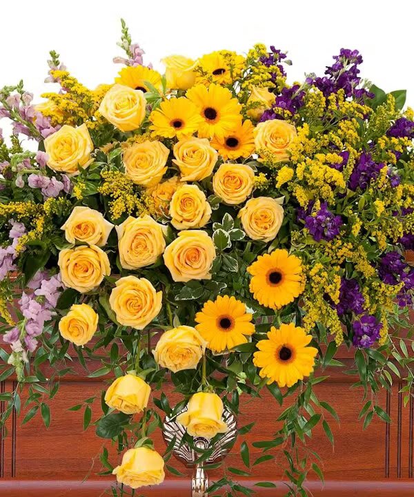 VIVID MEMORIES CASKET SPRAY - Bright, vivid yellow and purple flowers create a memorable casket spray.  The Vivid Memories Casket Spray is a radiant and expressive tribute, a flourish of color that celebrates a life lived with zest and passion. This striking spray combines the bright, optimistic tones of yellow with the regal and thoughtful hues of purple, creating a visual symphony that is as memorable as it is beautiful.  Yellow roses, the epitome of friendship and warmth, form the heart of the arrangement, their spiraling petals a sunburst of remembrance. Interspersed among them, the cheerful faces of sunflowers exude a sense of joy and nostalgia, reminiscent of sunny days and laughter. Solidago and other yellow accents add texture and depth, evoking the complexity and richness of the life being honored. Contrasting with the yellows, the purple flowers offer a touch of solemnity and dignity. They speak of the depth of affection and the respect held for the departed. The varied shades, from lavender to deeper purples, tell a story of individuality and uniqueness, a nod to the distinctive path the loved one walked.  Together, these flowers do not just adorn a final resting place; they proclaim a legacy of brightness and positivity, a lasting impression of a spirit that will continue to shine in the memories of those left behind. The Vivid Memories Casket Spray is not just a farewell; it's a celebration of a vivid life, forever etched in the hearts it touched.