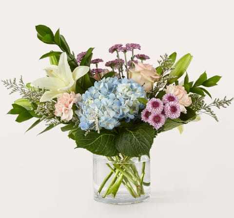 Slow Mornings Bouquet - Soft, serene, and effortlessly pretty. The Slow Mornings arrangement features peach carnations, blue hydrangea, white lilies, lavender button pompons, and fresh Israeli ruscus, styled low and lush in a clear glass cylinder.  The perfect pick-me-up for someone who could use a slower start to their day.  Approximately 14"H x 16"W.