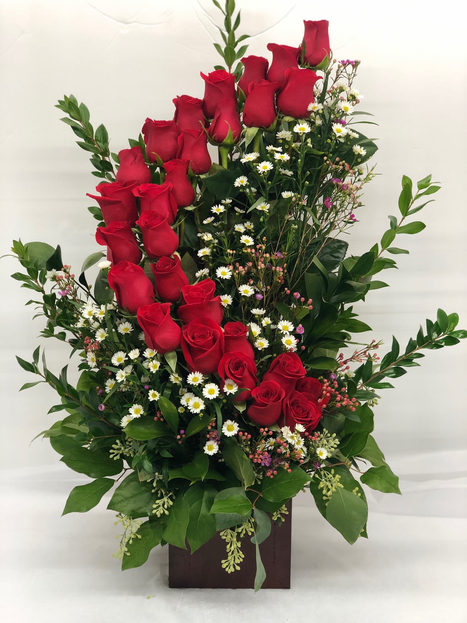 Tall arrangement of red roses with small white daisies in a dark box