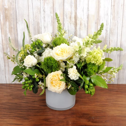 Low vase arrangement of cream roses, white flowers, and green accents in a frosted cylinder container.