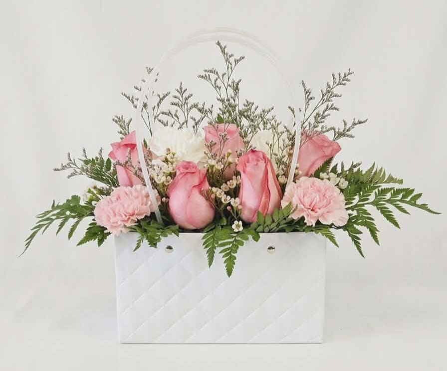 Blooming Floral Purse  - Soft pink roses and delicate pink-and-white carnations are arranged like a chic handbag bouquet in a white quilted gift bag-perfect as a birthday bouquet, a thank-you surprise, or a thinking-of-you arrangement. The Blooming Floral Purse offers a fresh, feminine presentation that reads as both playful and elegant, the kind of hand-delivered gift that makes the person you're thinking of feel seen and celebrated.  The arrangement combines fragrant roses and ruffled carnations with small white filler blooms and airy branching accents for texture, while fern-like greenery fans outward to create a full, balanced silhouette. The white quilted gift bag with matching white ribbon handles serves as both container and keepsake, giving the arrangement a polished, gift-ready look.  Ideal for friends, mothers, coworkers, or anyone who appreciates soft colors, this bouquet arrives ready to display.