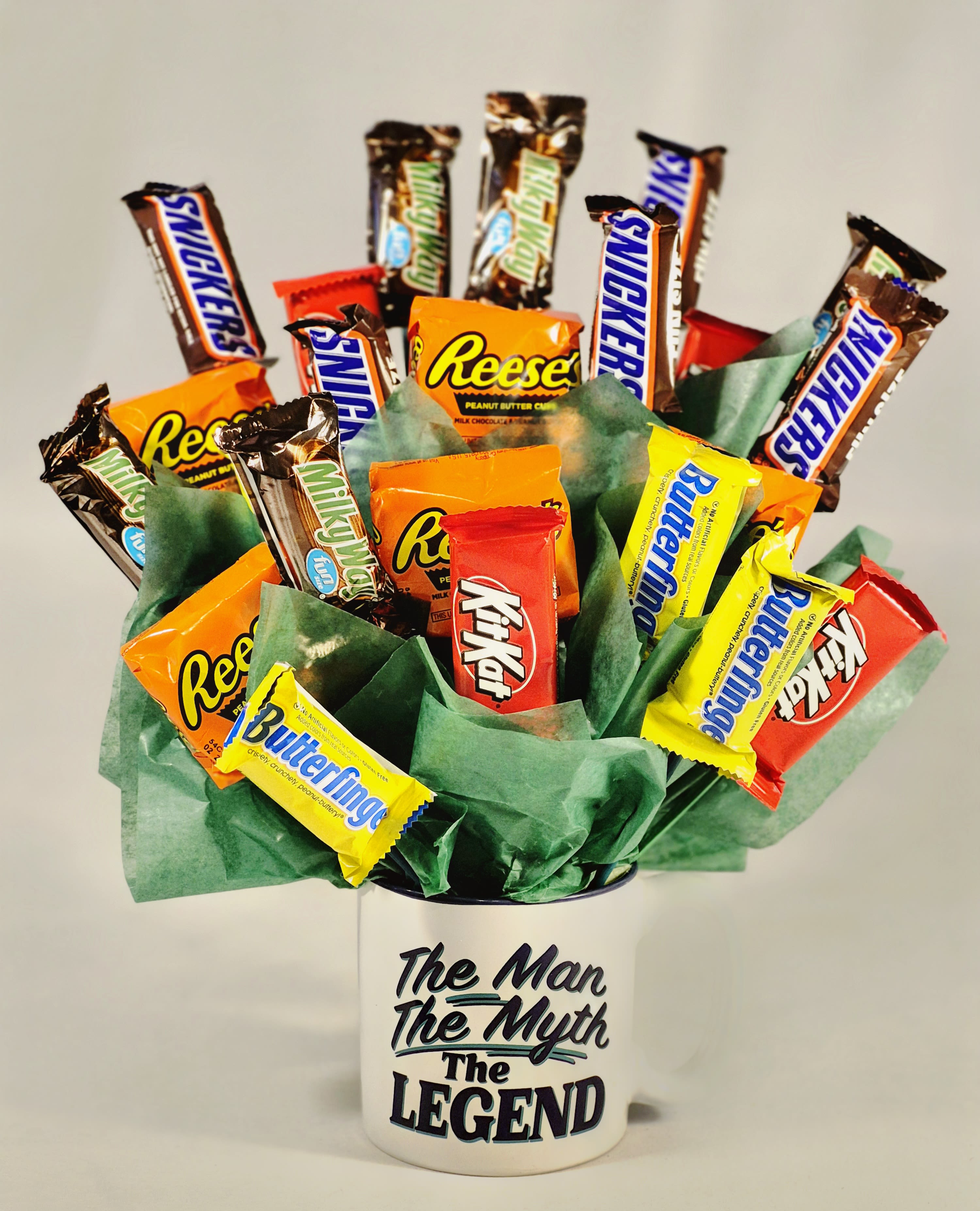 Man, Myth, Legend Father's Day Cup - Bright, playful, and built for Dad's big grin, this Father's Day cup puts sweet treats where a bouquet usually goes. A flower arrangement-style display of assorted snack-size candy bars-wrapped in bold oranges, yellows, reds and browns-burst from a white ceramic mug printed "The Man The Myth The Legend," making it an ideal Father's Day gift, birthday surprise, or casual congratulations present for the guy you want to celebrate.  The arrangement uses green tissue paper to add volume and a fresh pop of color around the base, letting the candy wrappers read like petals in a high-contrast, cheerful palette. The mug is food-grade and reusable, perfect for coffee, tools in the garage, or keepsake display. Candy varieties are assorted into a bouquet-style cluster so the recipient can pick favorites and enjoy a mix of textures and flavors.  Give this to the dad, coach, colleague, or friend who appreciates humor and sweets-it's the kind of gift that makes someone feel seen and celebrated without fuss. Easy to order and ready to give as-is, it arrives assembled and ready to brighten a morning routine or party table.
