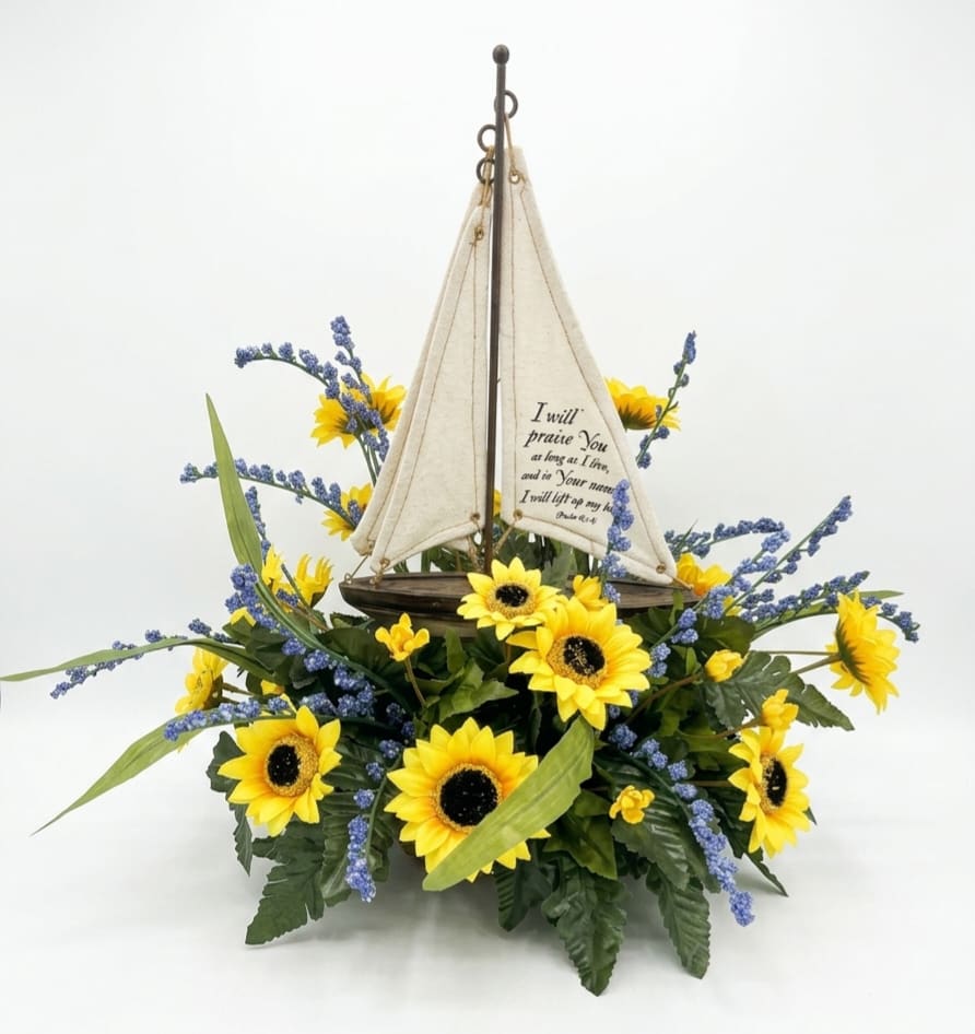 Psalm 63:4 Sailboat w/ Silk Arrangement  - Bright yellow sunflowers cluster around a handcrafted decorative sailboat to create a cheerful, nautical-themed flower arrangement perfect for everyday gifting or a thoughtful sympathy or encouragement piece. This flower arrangement and its cream-colored fabric sail - printed with Psalm 63:4 - makes a gentle, faith-forward statement that the person you're thinking of will notice and appreciate.   Blue filler blooms arc outward from dense green foliage to give the design movement and contrast against the sunny petals, while touches of white brighten the composition. The sailboat sits in the center of arrangement and the fabric sail gives a handcrafted, seasonal accent that pairs well with casual home décor or a bedside table.  Send this to celebrate a treasured friend, mark a small milestone, or offer comfort with a quiet scripture reminder; it's the kind of gift that feels both whimsical and sincere. Designed for display, the arrangement arrives ready to place where it can be enjoyed, and its fabric sail ensures the verse will remain a lasting keepsake. 15" T 12" W 