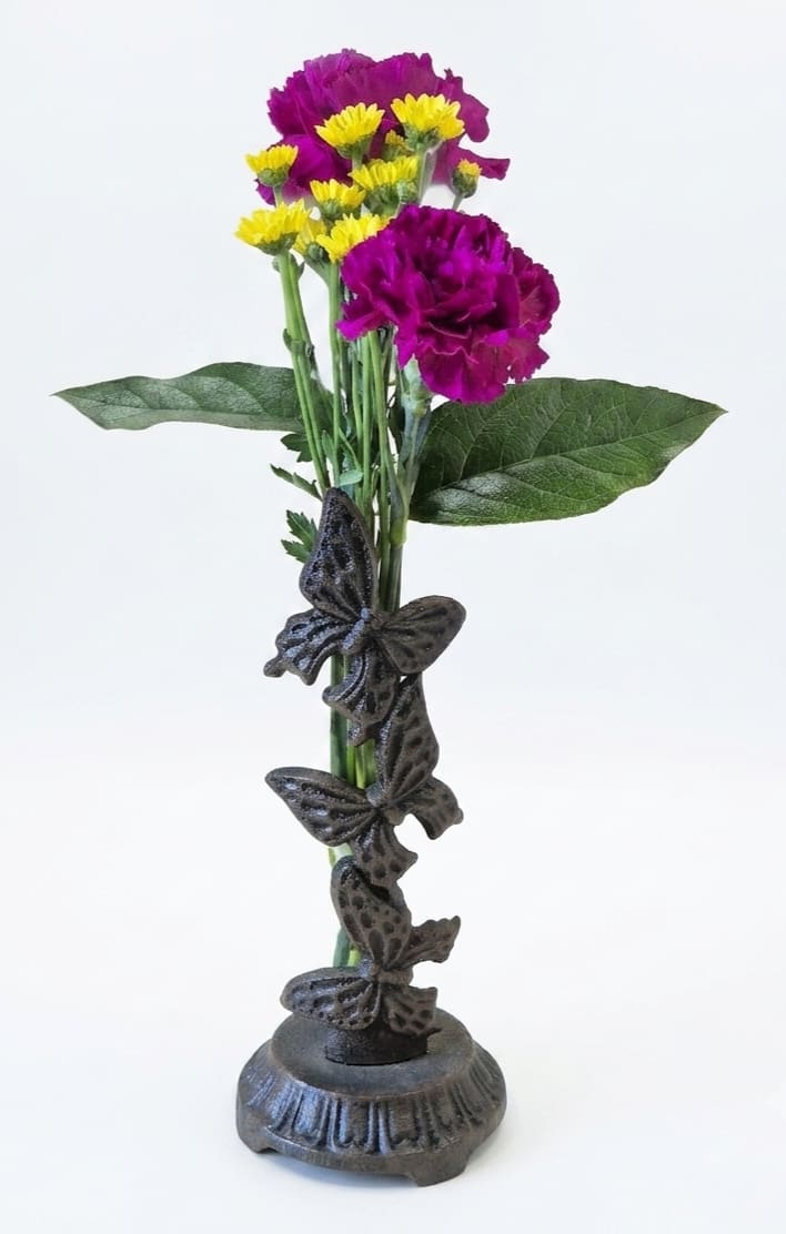 Rain Gauge Flower Bouquet - Vivid purple carnations and cheerful yellow daisy-like blooms create an eye-catching flower arrangement perfect for everyday display, birthdays, or a casual thank-you gift. This rain gauge flower bouquet pairs lively, textured blossoms with tall green stems to deliver a bright, uplifting presence the person you're thinking of will notice the moment it arrives.  The stems sit in a clear glass tube vase with measurement markings, mounted in a dark, textured metal stand and accented by a whimsical decorative butterfly wrapped around the stem area. The piece stands approximately 13" tall and doubles as a working rain gauge that measures up to 5 inches of rain - a charming, functional keepsake that brings both color and curiosity to a porch, windowsill, or entry table. Care: keep stems trimmed and water fresh for best vase life; the unit comes ready to display with its ceramic-like black textured container and glass tube insert.