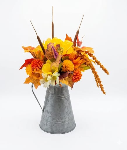 Pitcher of Fall Silk Floral - A cheerful harvest display in a galvanized metal pitcher, this fall silk floral arrangement is perfect for Thanksgiving decor or seasonal gifting. The compact 14" tall pitcher arrangement reads warm and welcoming-an autumn centerpiece or hostess gift that will make the person you're thinking of feel cozy and celebrated.  Dense clusters of silk chrysanthemums and daisies in bright yellow, orange, red, and white are layered with faux autumn leaves and tall cattail accents for height and texture. The rustic metal pitcher and decorative leaves give a country-chic, long-lasting look without the upkeep of fresh flowers.  Ideal for a Thanksgiving table, entryway, or as a mantel accent, this arrangement arrives ready to display-no watering or care needed-so it keeps its autumn charm throughout the season.