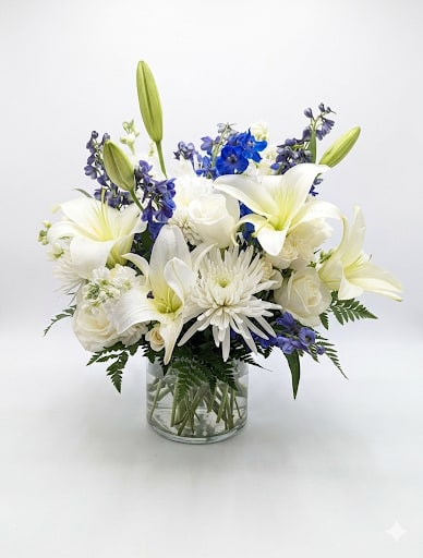 Thinking of You  - Pure white lilies and roses rise from a cloud of blue delphinium in this Thinking of You flower arrangement perfect as a sympathy arrangement, a thank-you gift, or a gentle way to celebrate someone's special day. The tall, graceful lily buds add movement and a hopeful lift so the recipient feels comforted and seen.  Arranged in a clear glass vase, the design is full and rounded with a clean, elegant palette of white, blue and green. The mix includes white roses, white lilies, white chrysanthemums (spider mums), blue delphinium and white stock, creating layered textures from the soft rose petals to the spiky delphinium and airy mums. At approximately 18" tall, it delivers presence without overpowering a bedside table, mantel, or office desk.  This is the kind of bouquet that quietly communicates care-whether you're sending condolences, gratitude, or warm thoughts.
